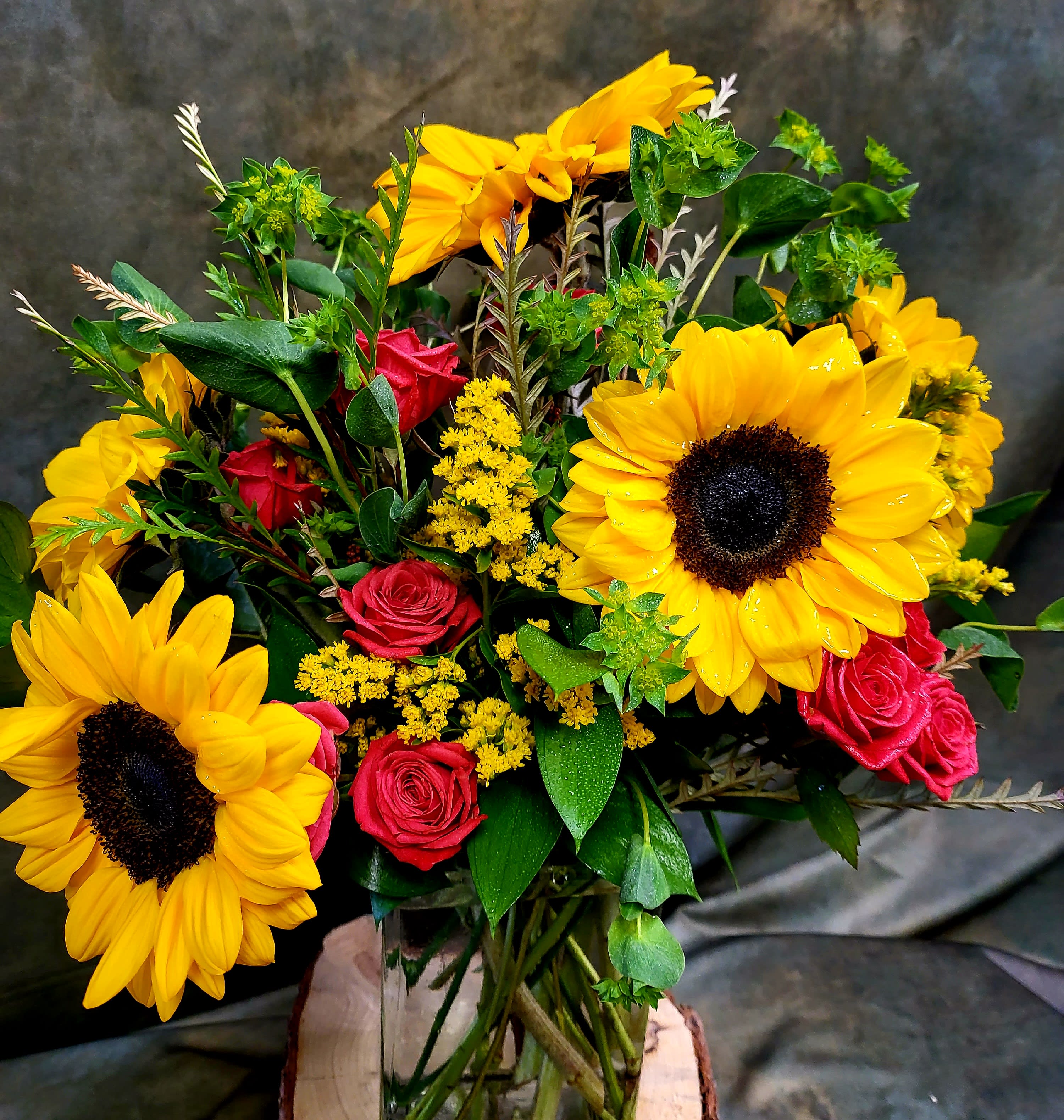 Simple Sunflowers  - Big, golden sunflowers steal the show in this Simple Sunflowers flower arrangement-a bright birthday bouquet or just-because gift that feels like a burst of sunshine for Mother's Day, Father's Day, anniversaries, or to cheer someone on while they recover. The warm contrast between large yellow sunflower heads and clusters of small red roses creates an uplifting, high-contrast look that reads as joyful and sincere to the person you're thinking of.  Fine yellow filler blooms are woven through the composition for added texture, and the stems are visible in a clear glass vase that sits on a natural wood slice base for a softly rustic finish. The mix of sunflower, rose, and greenery gives a generous, seasonal feel suitable for spring and summer celebrations or autumn and winter holidays-perfect for Thanksgiving tables, graduation gifts, or a cheery holiday centerpiece. Order with confidence for birthdays, get well wishes, thank-you gestures, or to say "I'm thinking of you."