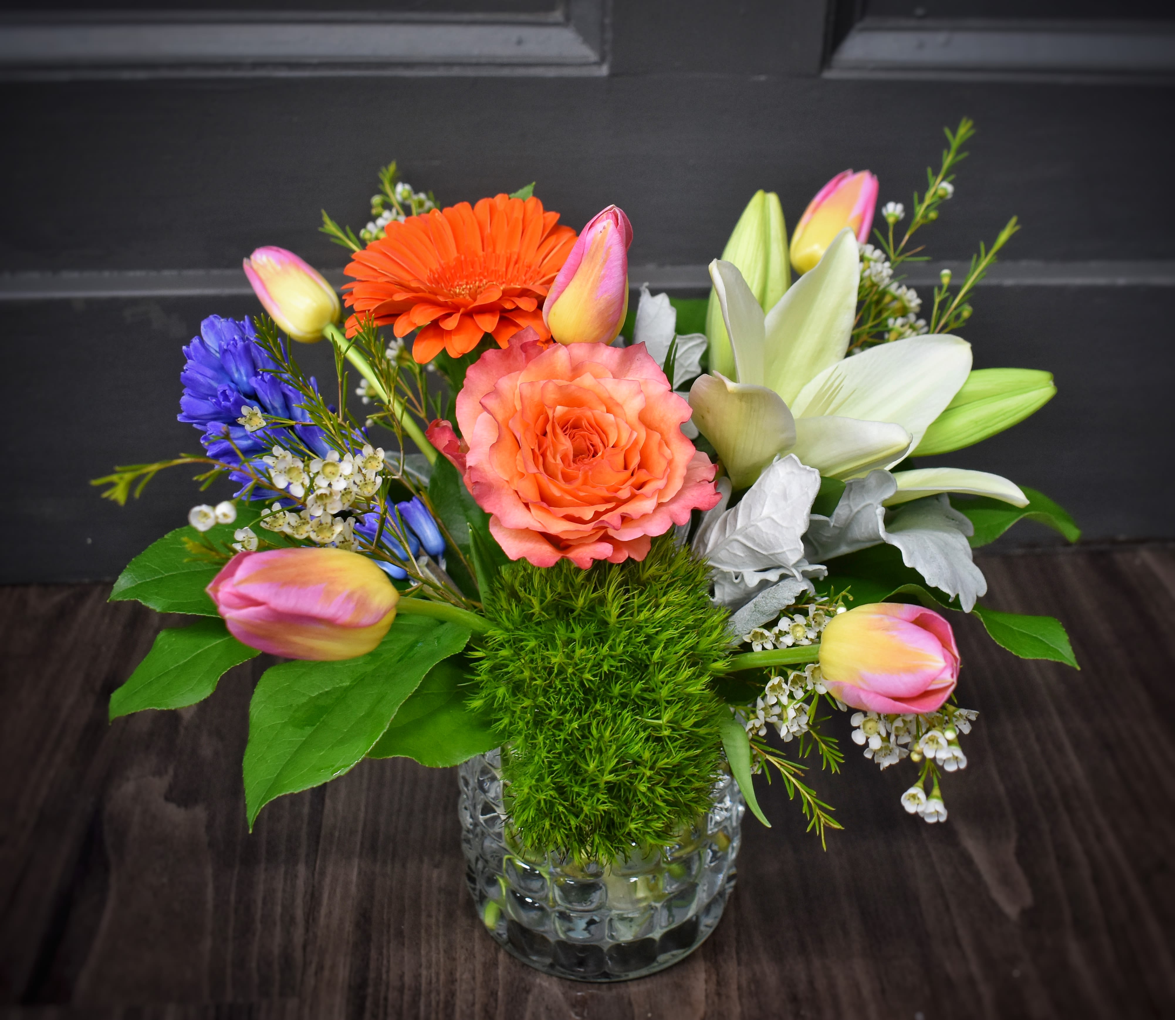 Symphony of Spring - A lively mix of coral, pink, orange and white blooms that reads like a garden in full swing - Symphony of Spring is a mixed-color flower arrangement perfect for a birthday, anniversary, or a bright "just because" surprise. The cheerful, asymmetrical composition and warm-cool palette make it especially welcome for spring occasions like Easter or Passover, and it's equally fitting to lift spirits as a get well gift.  This arrangement includes roses, spray roses, gerbera daisies, tulips, lilies, hyacinth, dianthus and small blue accent blooms arranged with airy white filler and a vivid green textured moss-like base. The flowers are presented in a clear glass vase that showcases the fresh stems; tones range from coral and peach to vivid orange, pink and pale whites, with silver-gray and green accents for contrast. The overall feel is full, garden-style, and fragrant where hyacinth and lilies are present.  Give this to the person you want to make feel noticed and uplifted - it reads joyful, celebratory, and thoughtful without being over the top. Available in a single vase size as shown; to keep blooms looking their best, refresh the water every two days and trim stems slightly when arranging the vase to prolong freshness.