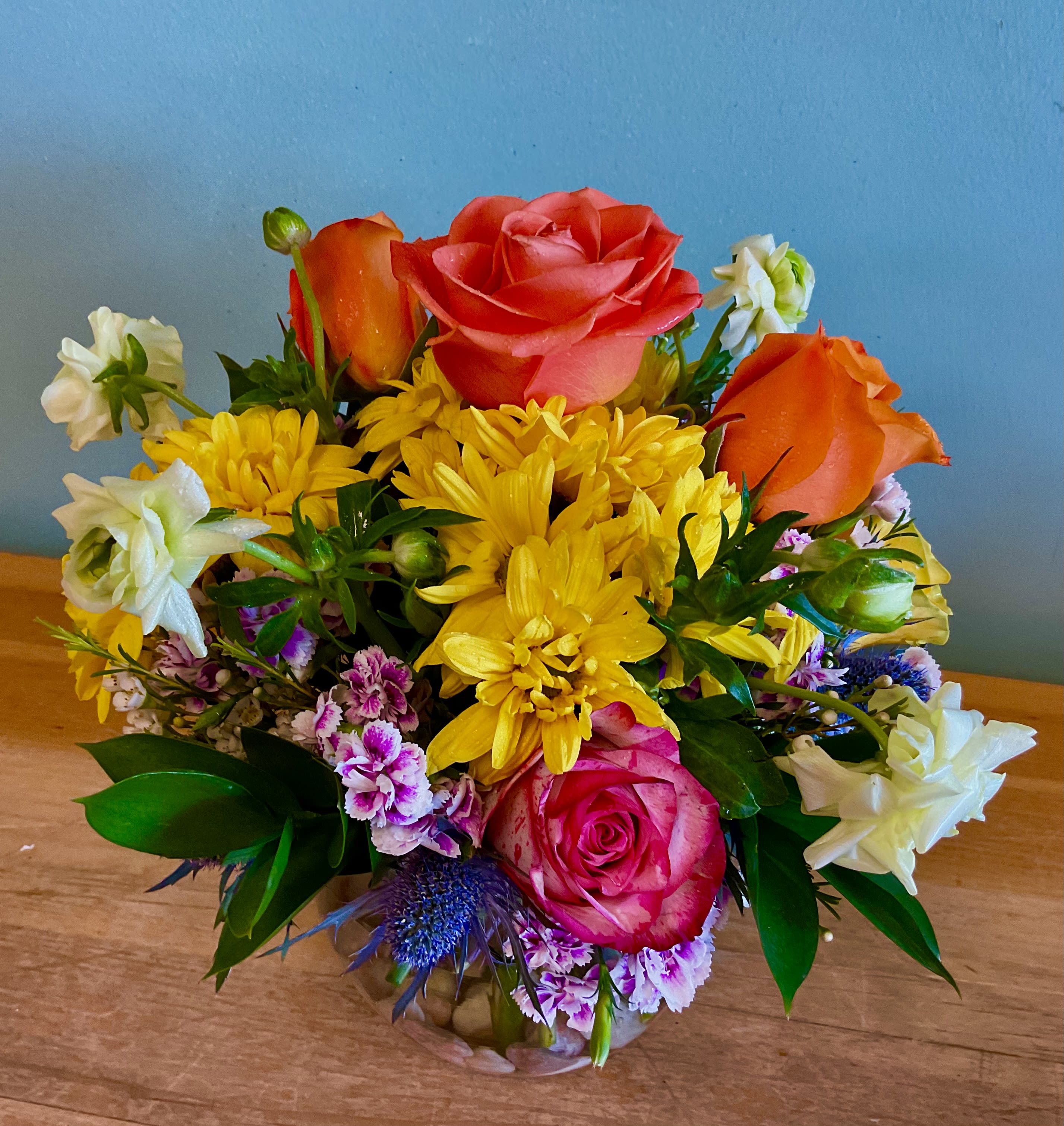 Bowl of Happiness  - A bright, garden-fresh bowl arrangement that feels like a pocket of sunlight-perfect as a Mother's Day gift, birthday bouquet, or just-because surprise. This rounded floral display mixes large pink and yellow roses with pale pink spray roses, cheerful white daisy-like asters, purple filler blooms, orange accents and soft carnations to create a lively, slightly romantic look that will make the person you're thinking of feel genuinely celebrated.  Arranged in a clear round glass vase, the composition is compact and full: taller focal roses rise above a dense middle layer of mixed bouquets and tiny accent flowers, while small white and yellow blooms soften the edges. Colors range from soft pastels to brighter jewel tones for a versatile palette that reads fresh in spring and summer but works year-round. Ideal for anniversaries, get-well wishes, graduations, housewarmings, thank-you gestures, new babies, or to say "I'm thinking of you," care is simple-keep water fresh and trim stems for lasting enjoyment.
