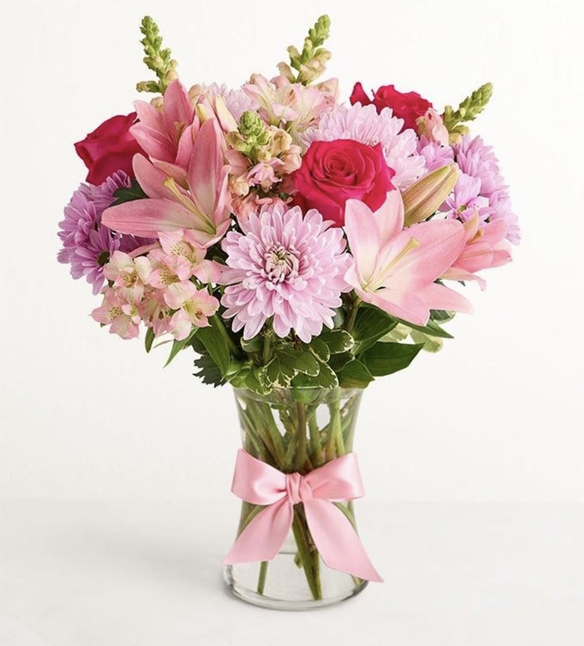 MIX FLOWERS GLASS /1  - A bright pink-and-red focal bloom greets the eye in this mixed flower arrangement, a cheerful flower arrangement perfect for birthday bouquets, anniversaries, or a just-because surprise. Pink lilies and roses anchor the composition so the person you're thinking of will feel truly celebrated, while softer blush chrysanthemums and white accents lend a gentle, romantic finish ideal for Valentine's Day or everyday gifting.  This full, rounded bouquet combines lilies, roses, chrysanthemums, alstroemeria, and snapdragons in shades of pink, red, white, and fresh green foliage, arranged in a clear glass vase and tied with a soft pink satin ribbon for a polished presentation. The layered textures-from the lilies' elegant petals to the delicate alstroemeria and vertical snapdragons-create visual depth and a lively silhouette that sits beautifully on a table or desk.  Made ready to display, the arrangement arrives hydrated in its vase so she can enjoy the color and scent right away; its balanced size works well as a centerpiece or a thoughtful bedside surprise. For best enjoyment, refresh the water and trim stems every few days to keep blooms looking their freshest.