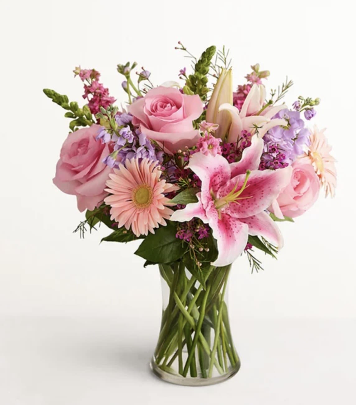MIX FLOWERS GLASS /2 - A romantic, pastel bouquet that reads like a whisper of affection - soft pink roses and a large pink-and-white lily take center stage in this flower arrangement for everyday gifting or a cheerful anniversary surprise. The mix flowers glass arrangement works beautifully as a birthday bouquet, a thinking-of-you gift, or a way to say congratulations without saying a word, giving the person you're thinking of an immediate feeling of warmth and care.  Inside a clear glass vase the blooms are arranged with long, visible stems for a fresh, airy look: pale pink gerbera daisy, fragrant stock, lavender and magenta filler clusters, and crisp white accents add depth and movement. The feminine palette of pinks, whites, purples, and green foliage feels both delicate and lively, creating a textured, garden-fresh impression that delights at first glance.  Perfect for brightening a bedside table, desk, or entryway, this bouquet is the kind of gift that makes someone feel truly seen and cherished. Vase is included; stems are shown through the water for an elegant, natural presentation - no extra styling required.