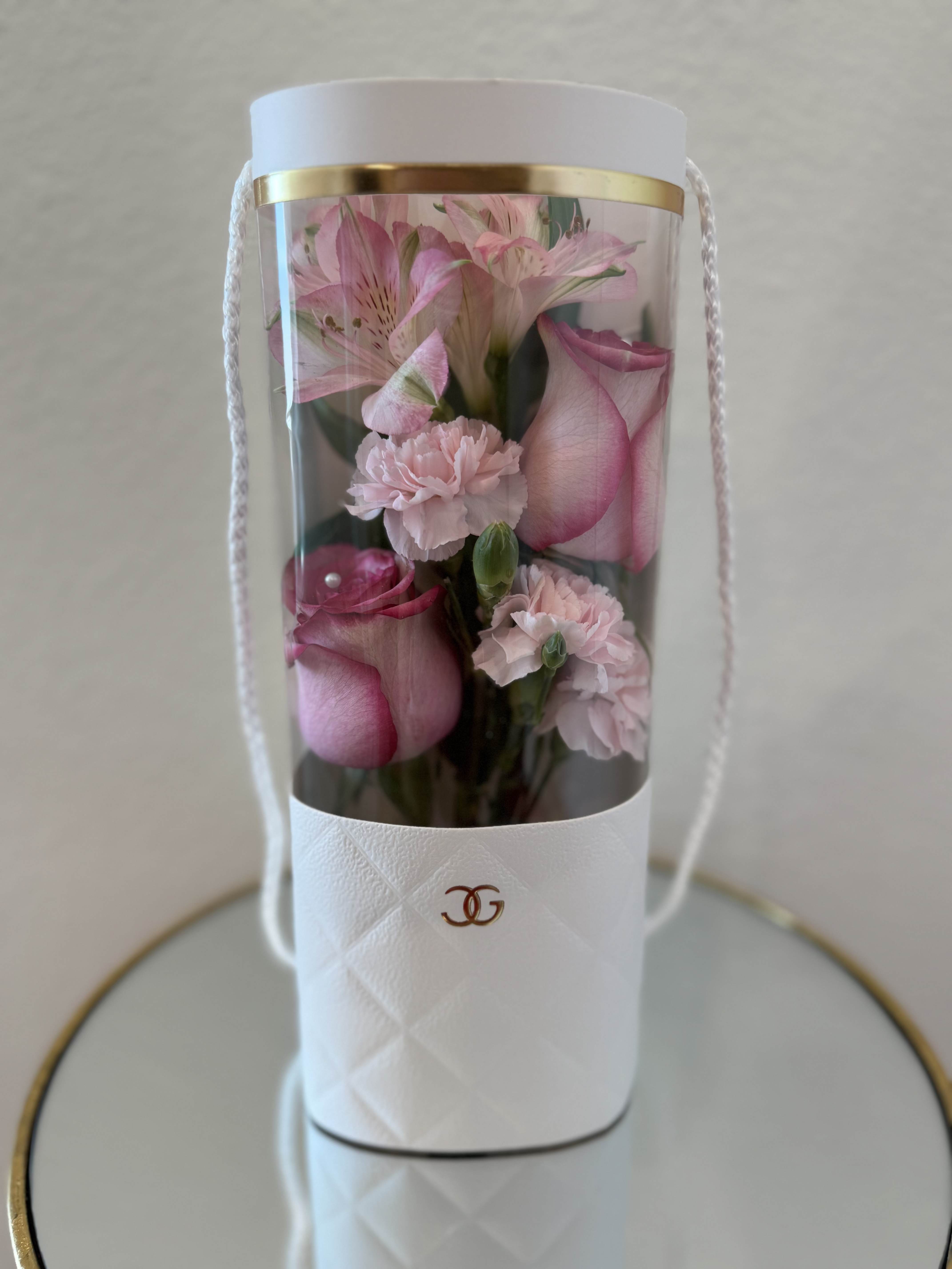 WITHE TALL BAG - Soft pink roses and pale carnations rise from a tall white cylindrical gift box to create a compact, romantic flower arrangement perfect for anniversary or birthday bouquets and thoughtful thank-you gifts. The clear front cover and neat vertical presentation let the recipient see the delicate blooms while preserving their freshness, while gold trim and rope handles give the piece a polished, gift-ready look that reads as both elegant and intimate.  Inside the white box the roses and carnation clusters are tightly grouped with touches of green and a few lily-like blooms for height and subtle contrast, all arranged to emphasize texture and a soft, blushing palette. The clear plastic cover protects the stems and keeps the bouquet feeling refined; the box doubles as a keepsake container so the presentation lasts beyond the flowers themselves. This is the kind of arrangement that makes someone feel seen and celebrated-perfect for sending love, saying thank you, or marking a special birthday.