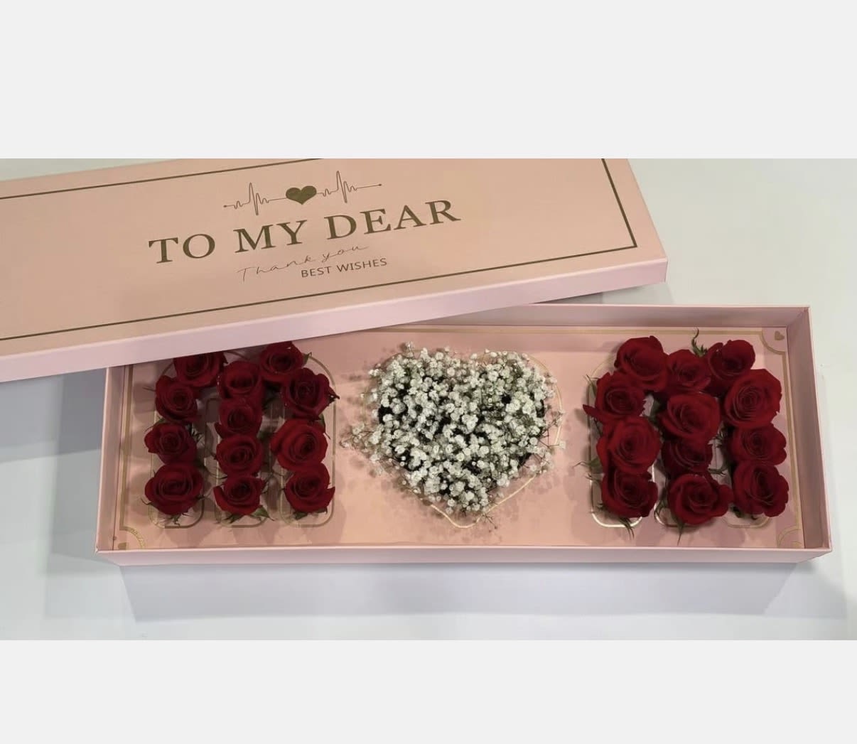 PINK MOM BOX  - A long pale-pink gift box filled with three lush groupings of deep red roses creates an immediate wow - a Mother's Day or anniversary flower arrangement that reads as both romantic and celebratory. Two blocks of velvety red roses frame a central heart-shaped cluster of white baby's breath, so the person you're thinking of will feel seen and cherished the moment they lift the lid. The pink presentation box and matching lid are printed with a sentimental message, making this a keepsake-ready gift.  Tucked among the roses are delicate butterfly accents, a gold "Happy Mother's Day" topper, and a neat gold topper for added sparkle, while a selection of chocolate truffles adds a delicious extra treat. The tightly packed blooms give a full, luxurious appearance; colors are deep red roses, crisp white baby's breath, and soft pink packaging for a polished contrast. Size and care follow standard boxed-arrangement guidance: keep in a cool spot out of direct sun and add water according to the included instructions to preserve freshness.