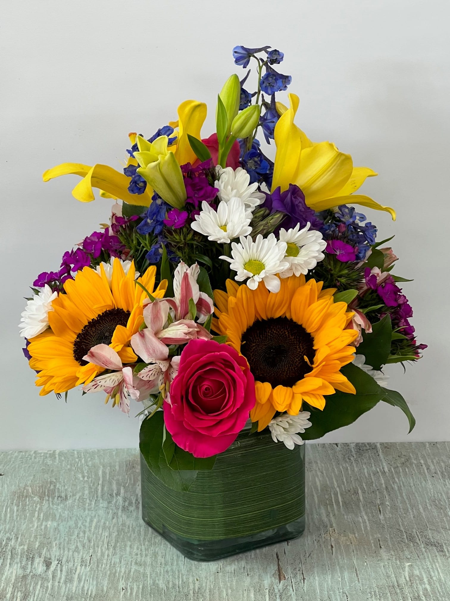 Colorful mixed bouquet in a square glass vase with sunflowers, lilies, roses, and daisies