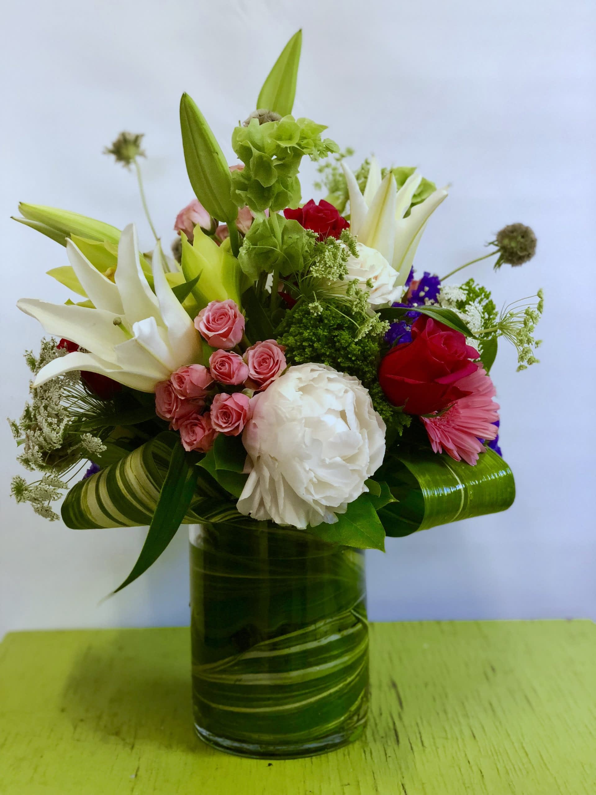Mixed bouquet in a green glass vase with lilies, roses, and a white peony