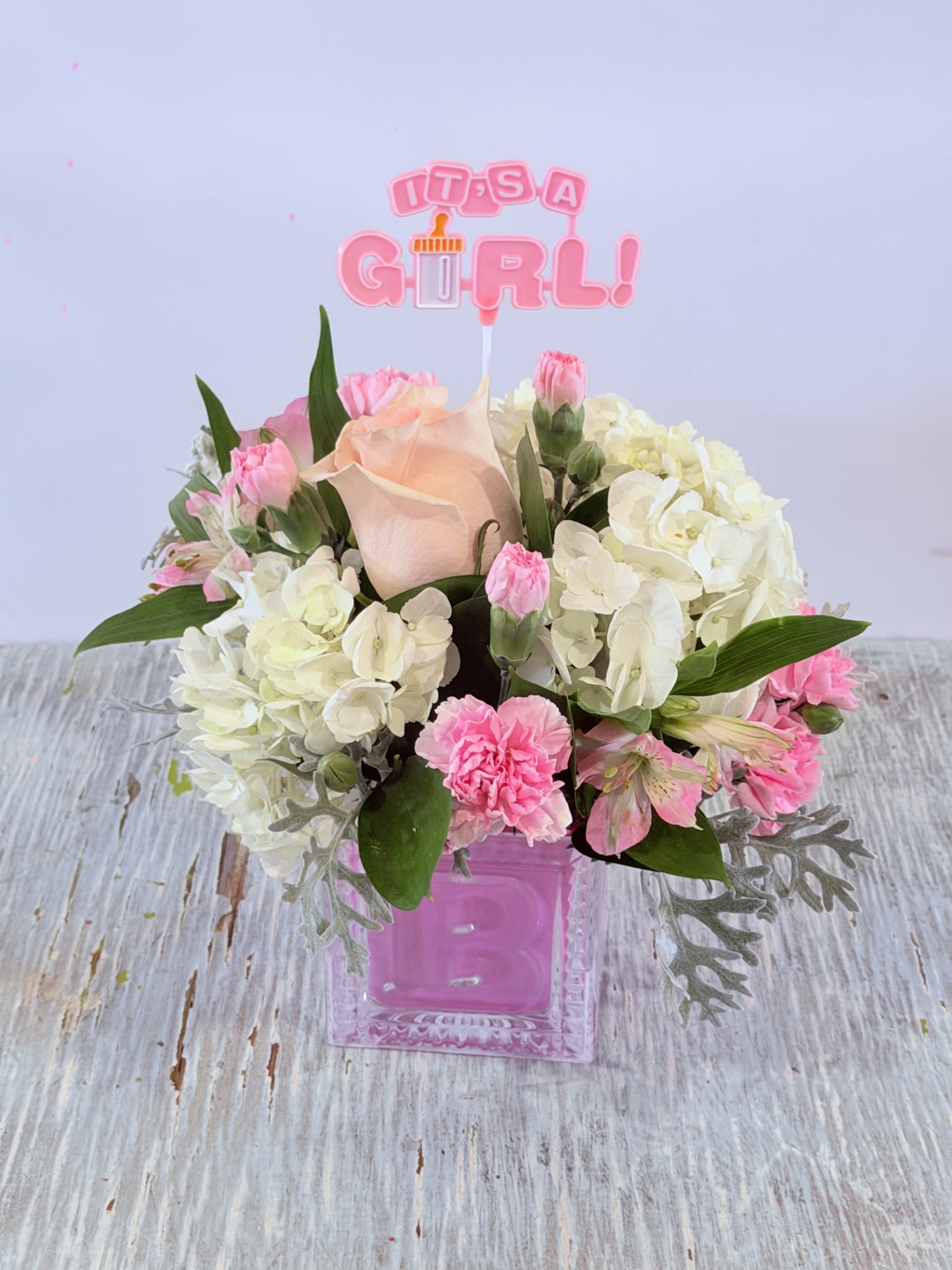 Welcome Baby Girl  - Pale blush roses paired with cloudlike white hydrangeas and soft pink carnations create a tender, celebratory bouquet perfect as a welcome baby girl gift. This baby-themed arrangement reads gentle and feminine the moment she sees it-an "It's a Girl!" topper and a glossy pink glass vase make the occasion unmistakable for a newborn celebration.  Compact and rounded, the bouquet also features white and pink alstroemeria, soft green foliage, and silvery filler that frames the blooms for a delicate, textured look. The pieces are arranged close together for a lush, full presentation that fits beautifully on a bedside table or nursery dresser.  Give this to new moms, grandparents, or the whole family to announce your joy-the arrangement is designed to make her feel warmly celebrated and seen. Vase included; simple care is to keep water fresh and trim stems every few days to keep the hydrangeas and roses at their best.