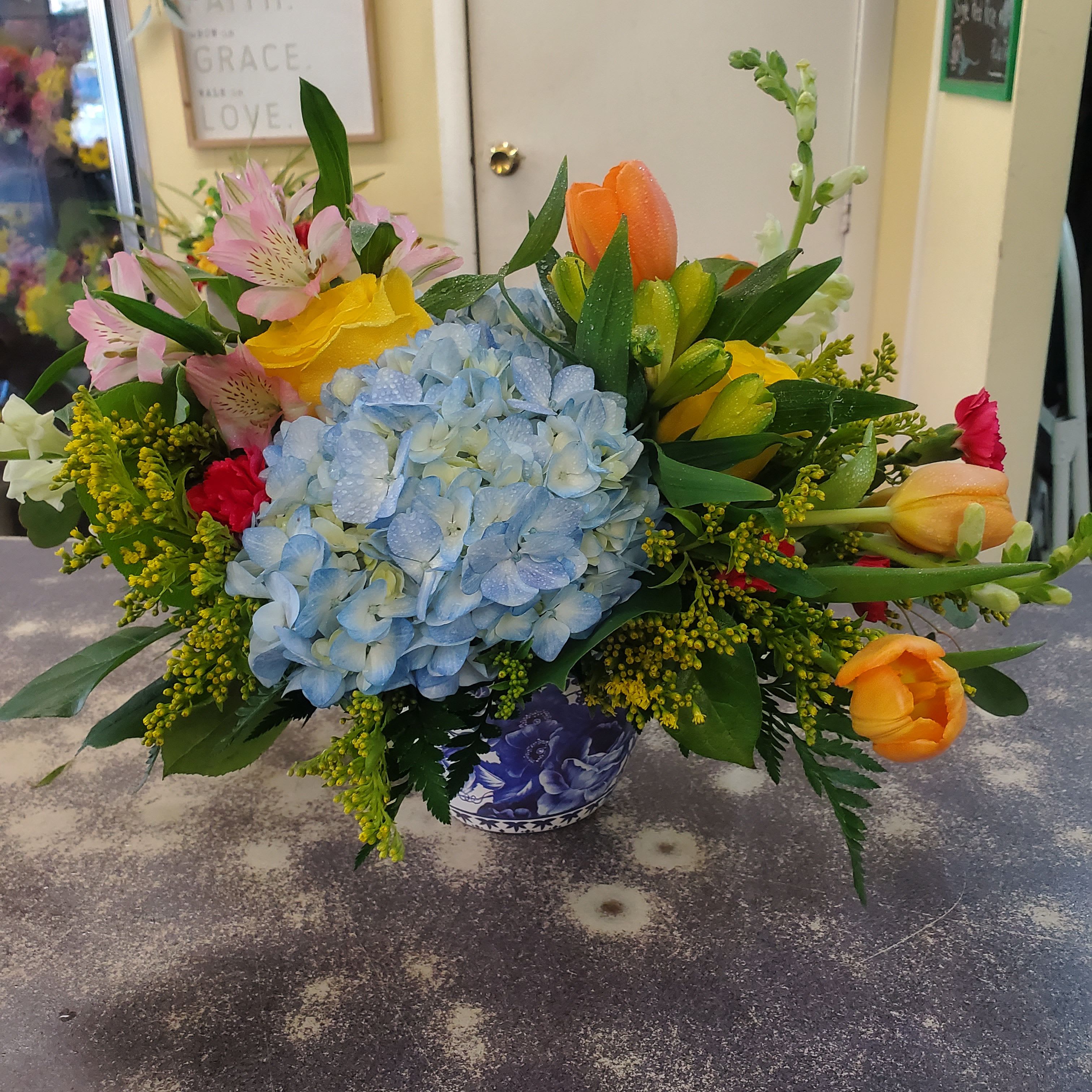  Blue Skies Bouquet  - Arrangement designed in a blue and white ceramic container with assorted blues, yellows and orange flowers accented with assorted natural greens.  **Low compact design **