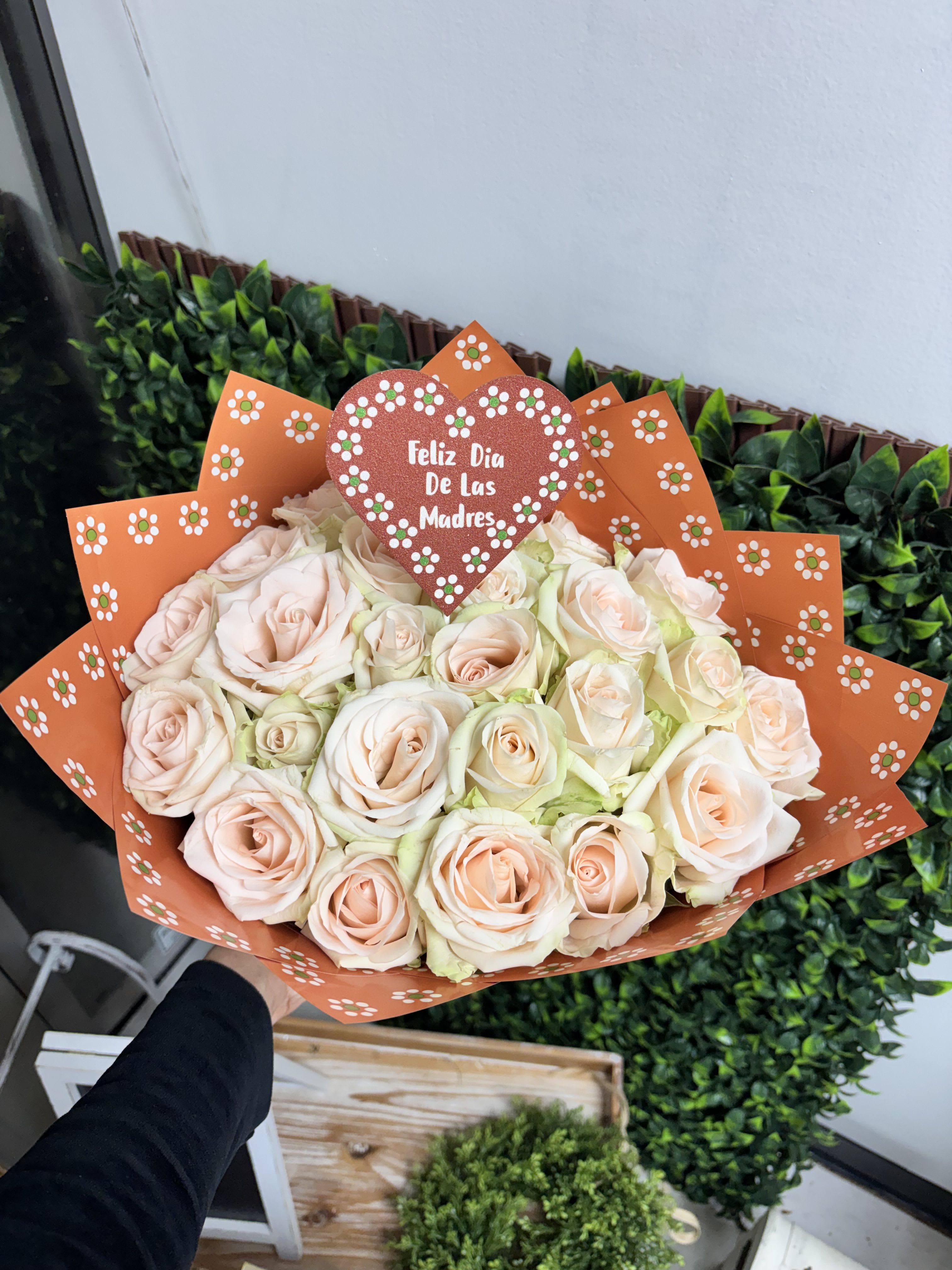 Feliz Dia De Las Madres 24 roses  - Pale peach and blush roses gathered into a lush, rounded hand-tied bouquet make an instantly warm Mother's Day gift that reads as both tender and celebratory. This 24-rose arrangement is designed for "Feliz Día de las Madres"-the glittery heart topper says it plainly-so the person you're thinking of will feel honored and loved the moment they see it. The soft pink, peach, white, and subtle orange rose tones create a gentle, approachable palette that works equally well for a mother, grandmother, or the special woman in your life.  Every stem is clustered tightly for a full, cushiony silhouette and wrapped in bright orange decorative paper printed with small floral motifs, giving the bouquet a lively pop that contrasts beautifully with the muted roses. The heart-shaped topper adds a festive, keepsake element without overpowering the blooms. This is the kind of bouquet that feels thoughtful for Mother's Day, anniversaries, or to say "gracias"-she'll feel genuinely celebrated. Arrange arrives hand-tied and ready to place in a vase; keep roses in fresh water and away from direct heat to extend their bloom.