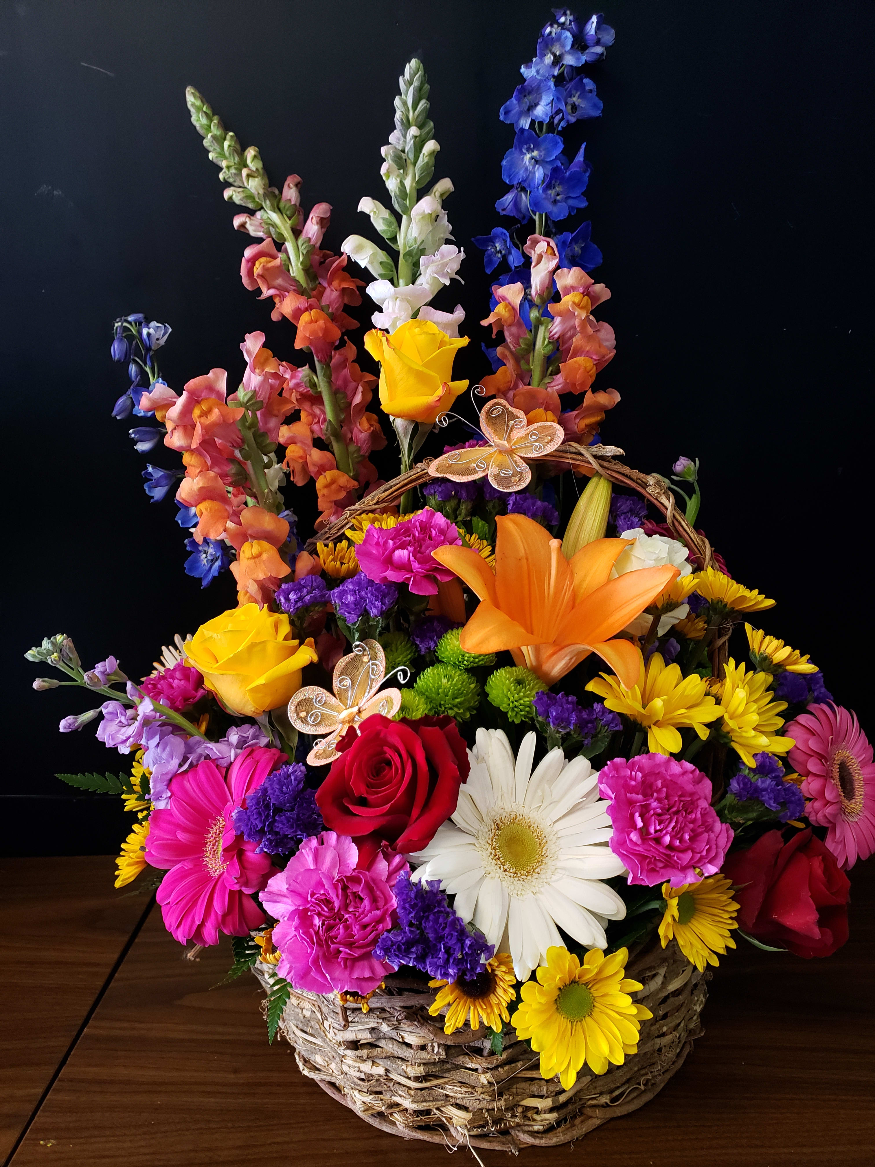 My Mad Maddy  - Bright, garden-fresh color takes center stage in this cheerful basket arrangement-perfect as a birthday bouquet, a vibrant Mother's Day or graduation gift, or a "get well soon" pick-me-up. The tall snapdragons and delphinium create a graceful, airy backdrop while the saturated band of red and yellow roses, magenta carnations, and sunny mums form a joyful focal point that makes the recipient feel truly celebrated.  This full, low-to-tall design is gathered in a woven white wicker basket and includes sunflowers, gerbera daisies, lilies, stock, chrysanthemums, button mums and mixed accent blooms for a lively, mixed-color palette of purple, yellow, red, pink, orange and blue. Decorative metal and iridescent butterfly picks are tucked among the stems for a whimsical finishing touch. Sized to fill a room, it's an easy choice for occasions from anniversaries, Housewarming and Thanksgiving to Sympathy, holiday celebrations, or simply saying "Thinking of You"-arrives ready to display with simple vase-care instructions included.