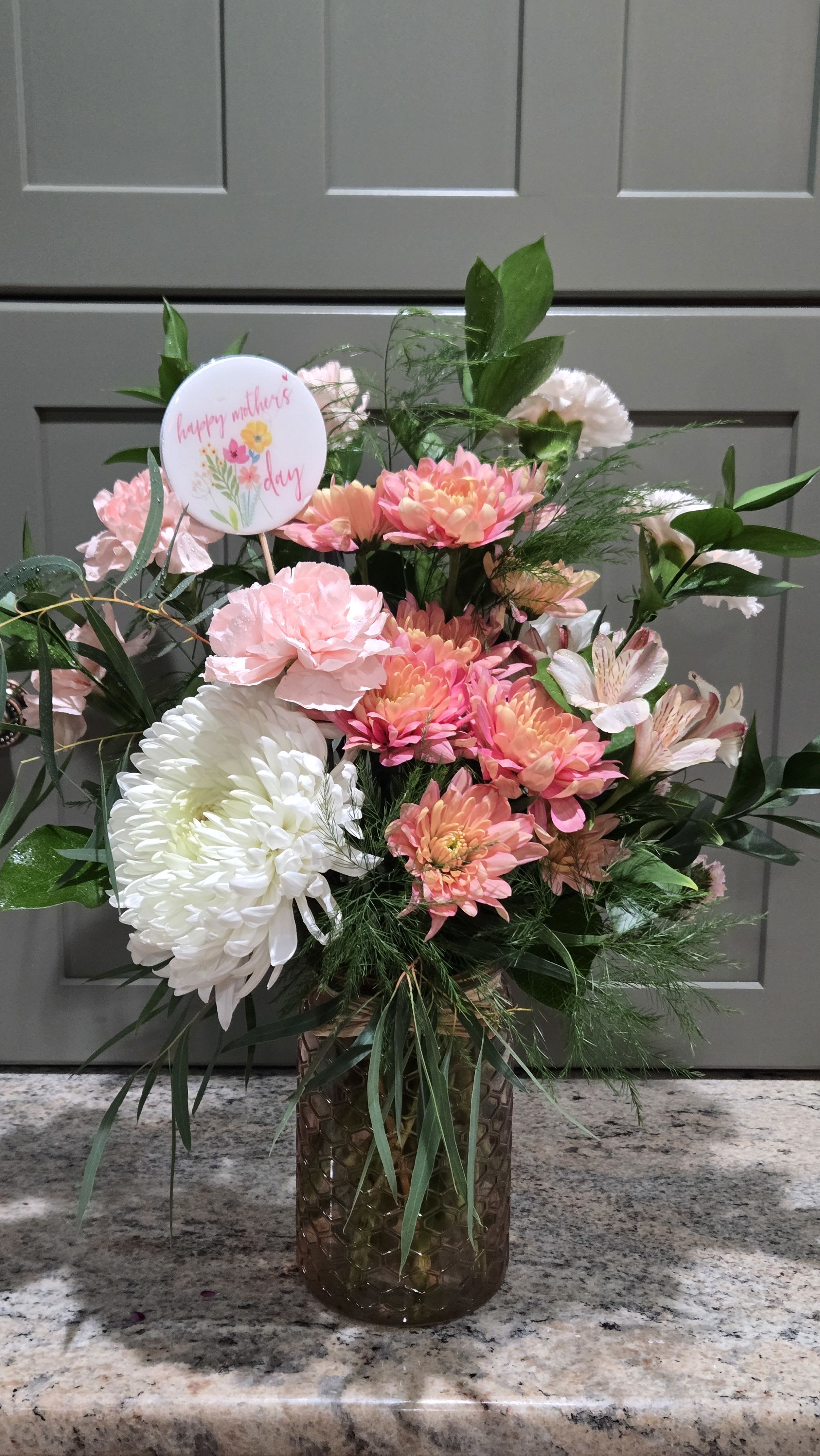 Peachy Momma - Soft peach and pink blooms gathered in a textured glass vase create an instantly cheerful Mother's Day flower arrangement that feels both celebratory and heartfelt. This flower arrangement for Mother's Day features large chrysanthemum heads alongside ruffled carnations, alstroemeria, and daisy-like blooms so the person you're thinking of will feel noticed and adored. The round "Happy Mothers Day" pick makes the gift unmistakably for Mom while keeping the mood light and feminine.  Creamy whites, peach, and a range of pink tones are layered with airy greenery for a garden-style, rounded silhouette that looks just-cut and fresh. The arrangement sits in a textured glass vase and includes a decorative round sign on a stick; small yellow accents in the sign add a sunny contrast to the predominantly pink and white palette. The compact, closely packed design offers good presence on a table or counter without overwhelming a small space.  Perfect for Mother's Day, a springtime thank-you, or to mark a special birthday, Peachy Momma reads as thoughtful and joyful to the recipient. Arrange for local delivery from Carroll's Florist so the flowers arrive hydrated and ready to display; its classic shape and durable vase also make it easy to refresh the water and enjoy for days.