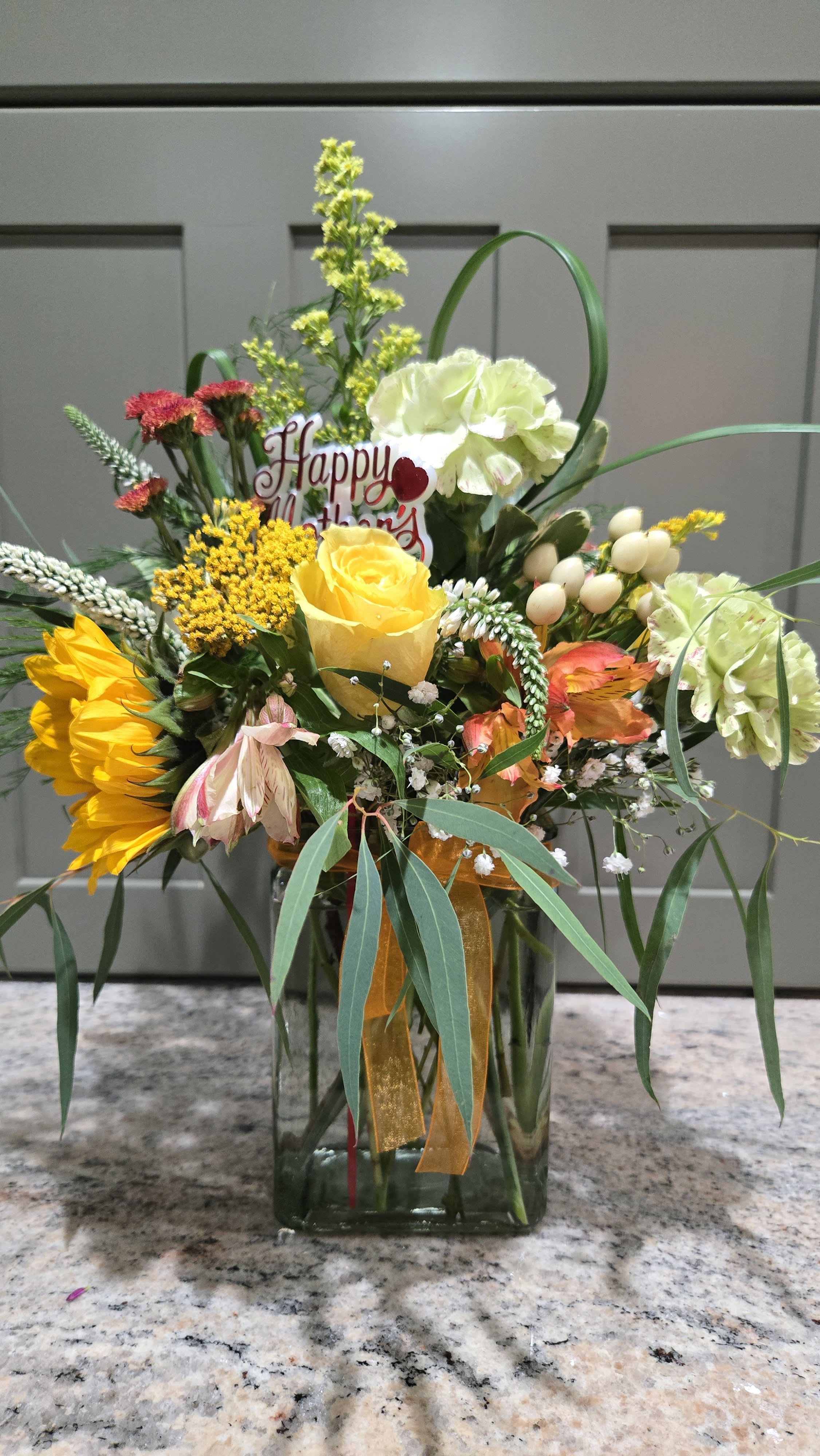 Sunshine For Mom - A bright yellow rose takes center stage in this cheerful Mother's Day flower arrangement, surrounded by sunflowers, orange and white carnations, pink alstroemeria and sweet daisy-like filler blooms-perfect as a Mother's Day bouquet or a celebratory gift to make her smile. The mix of sunny yellow, warm orange, soft white and pink creates an uplifting, joyful impression that says "you are loved" the moment she sees it.  Arranged in a clear rectangular glass vase, the design features long, arching foliage for movement and a sheer orange ribbon for a festive touch. A small "Happy Mother's Day" pick is included to make the occasion unmistakable. The compact vase keeps the bouquet table-friendly while showcasing the blooms' colors and textures-roses and sunflowers for bold focal points, alstroemeria and daisies for delicate contrast.  This is the kind of gift that will make Mom feel truly celebrated-bright, warm and full of life. Order with confidence for same-week delivery and follow simple care: keep water fresh, recut stems every few days, and display in a cool spot away from direct heat to extend enjoyment.