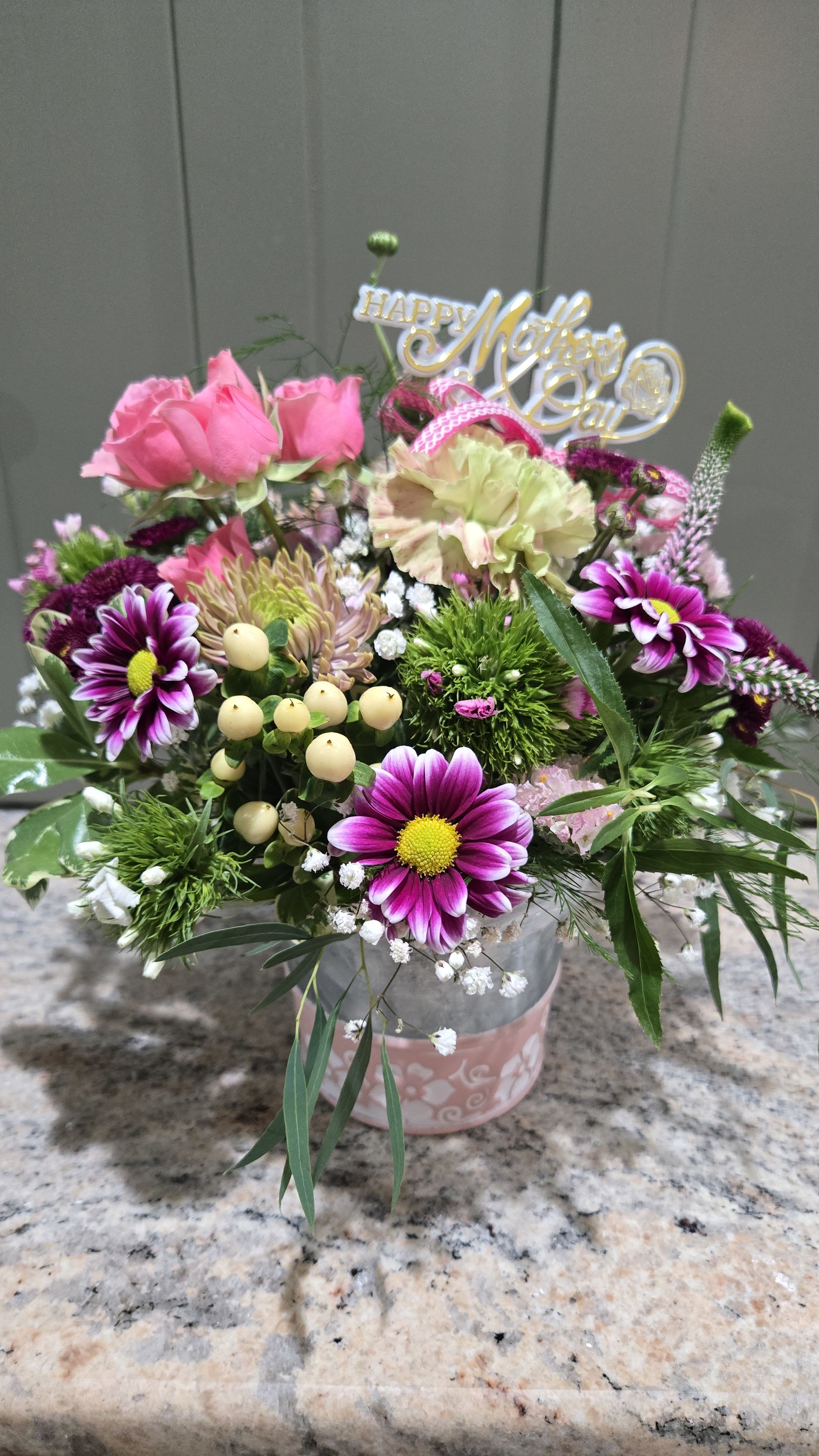 Moms Are Everything  - A joyful Mother's Day flower arrangement that feels like a warm hug - perfect as a Mother's Day bouquet or a pick-me-up for the mom who does it all. This compact flower arrangement blends pink roses and pale carnations with purple-and-white daisy chrysanthemums and creamy filler blooms to create a layered, feminine palette of magenta, blush, white and fresh green foliage. The gold-and-white "Happy Mother's Day" topper gives it an unmistakable, celebratory message she'll see the moment the box opens.  Arranged in a simple ceramic vase and finished with a pink ribbon and decorative topper, the mix reads lively and hand-crafted without taking up a lot of space-ideal for a kitchen table, bedside dresser, or office desk. The combination of roses, carnations, daisies and chrysanthemums provides a variety of textures and gentle fragrance that will make her feel truly celebrated; this is the kind of bouquet that says "thank you" and "I love you" in one cheerful gift.