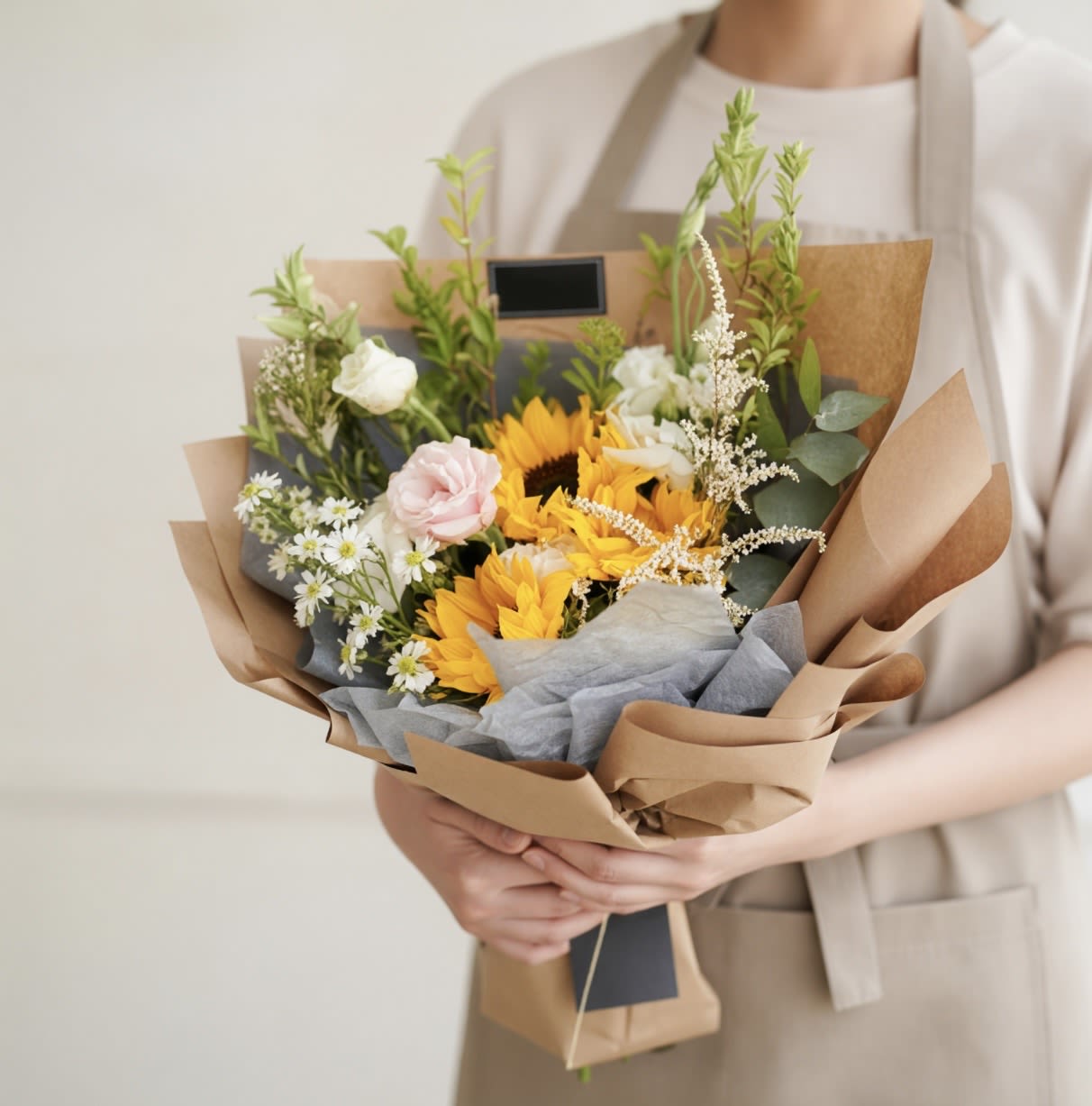 Golden Bloom Graduation Bouquet (Seasonal Blooms) - Celebrate their milestone with a bouquet as bright as their future. The Golden Bloom Graduation Bouquet features a cheerful mix of seasonal blooms—highlighted with radiant sunflowers, soft garden accents, and lush greenery—beautifully wrapped in our signature kraft and tissue layering.  Designed to make a statement for photos, gifting, and unforgettable moments, this bouquet is perfect for honoring your graduate in style.  Details:  Handcrafted with fresh, seasonal flowers Wrapped bouquet (no vase) for easy carrying at ceremonies Color palette may vary slightly based on availability Designed in our signature airy, garden-style aesthetic   Perfect for graduations, celebrations, and proud moments worth remembering.