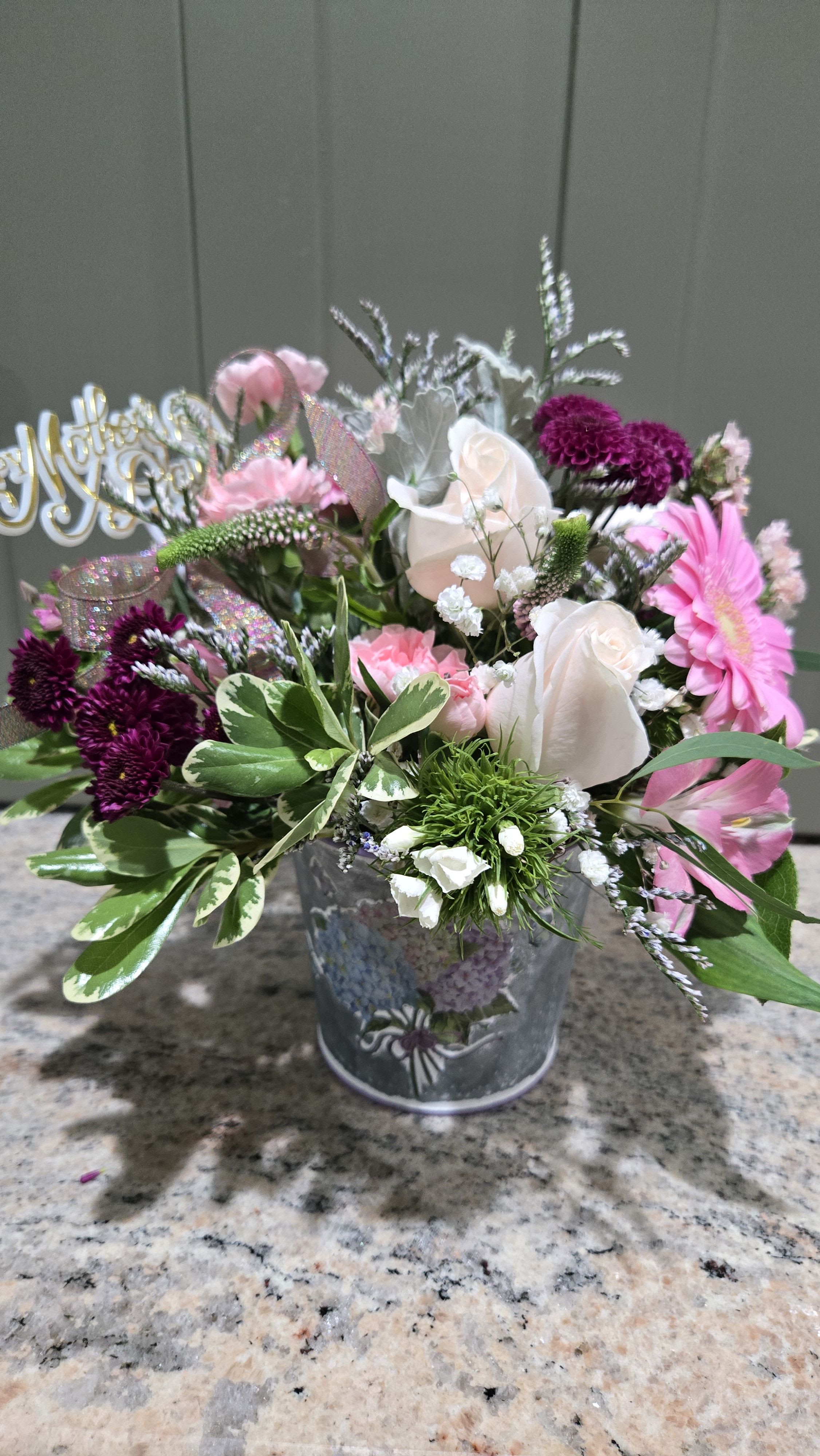 Mum's Love - Pale pink roses and bright pink gerbera daisies meet deep purple chrysanthemums in a charming hand-gathered display designed to make mom feel seen and celebrated. This flower arrangement for Mother's Day (also perfect for birthdays, anniversaries, or just because) balances soft pastels with jewel-toned accents so the recipient will feel both cherished and uplifted the moment they see it.  Roses, carnations, gerbera daisies, chrysanthemums and small white filler flowers are layered with varied greenery for a full, textured silhouette. The blooms are arranged in a galvanized-style metal bucket, tied with a glittery decorative ribbon and an iridescent looped bow; a metallic script pick and a gold-and-white "Happy Mother's Day" topper add a handcrafted, festive touch. The mix of lavender, purple, pink, white and green creates a fresh spring palette that also reads romantic enough for love and anniversary gestures or gentle enough for a new baby, thank you, holiday, or thinking-of-you gift.  This cheerful, slightly rustic presentation is sized to sit beautifully on a table, counter, or bedside - the kind of bouquet that brings warmth to everyday moments and makes special occasions feel intimate. Order with same-week delivery when available to ensure peak freshness and the fullest impact for the person you're celebrating.