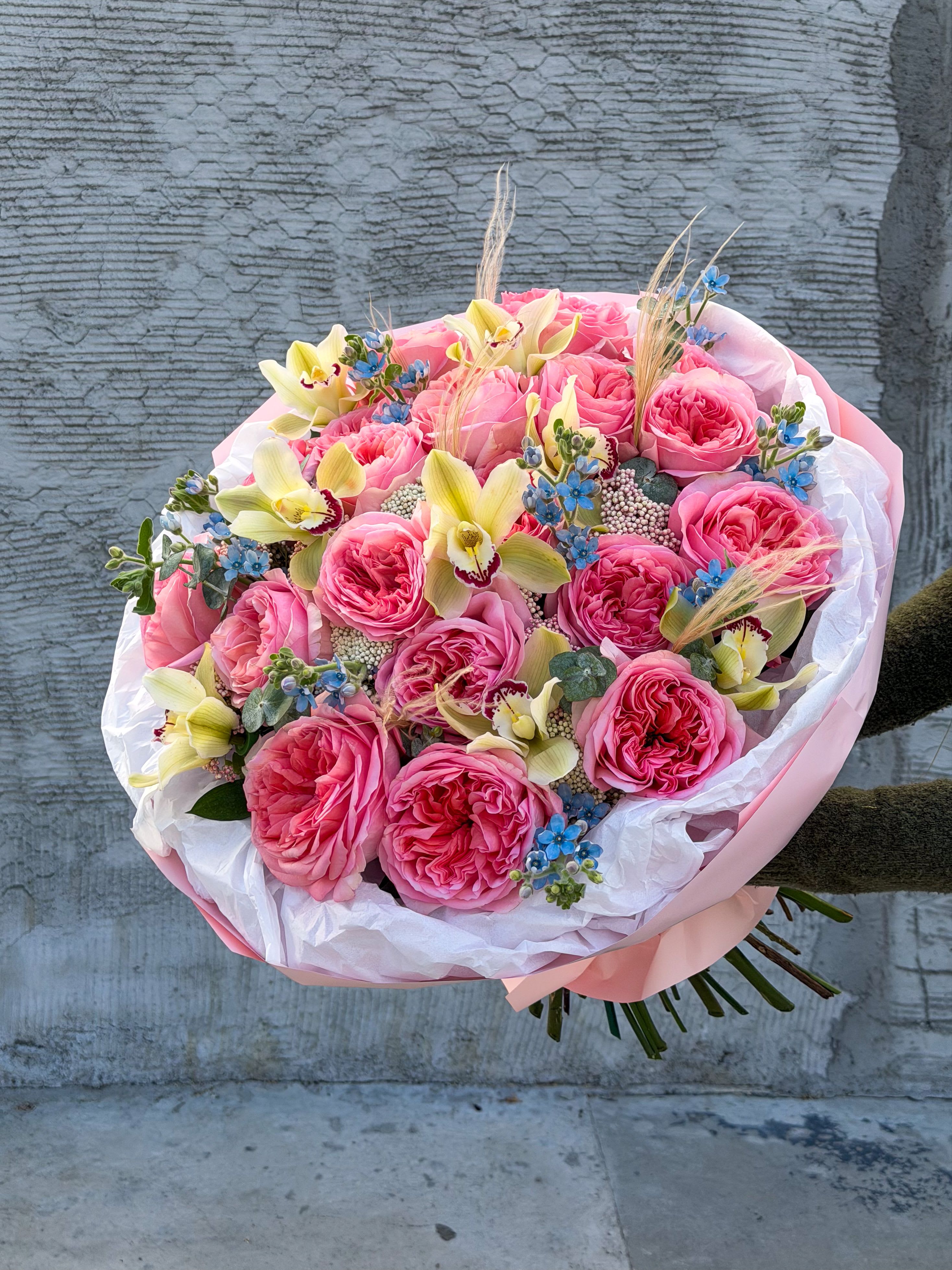 Julia - A soft, romantic composition built around lush pink garden roses, designed to feel feminine, elegant, and full of quiet confidence. Accented with delicate cymbidium orchids, airy blue details, and textured elements, this bouquet has that signature “expensive but effortless” look.