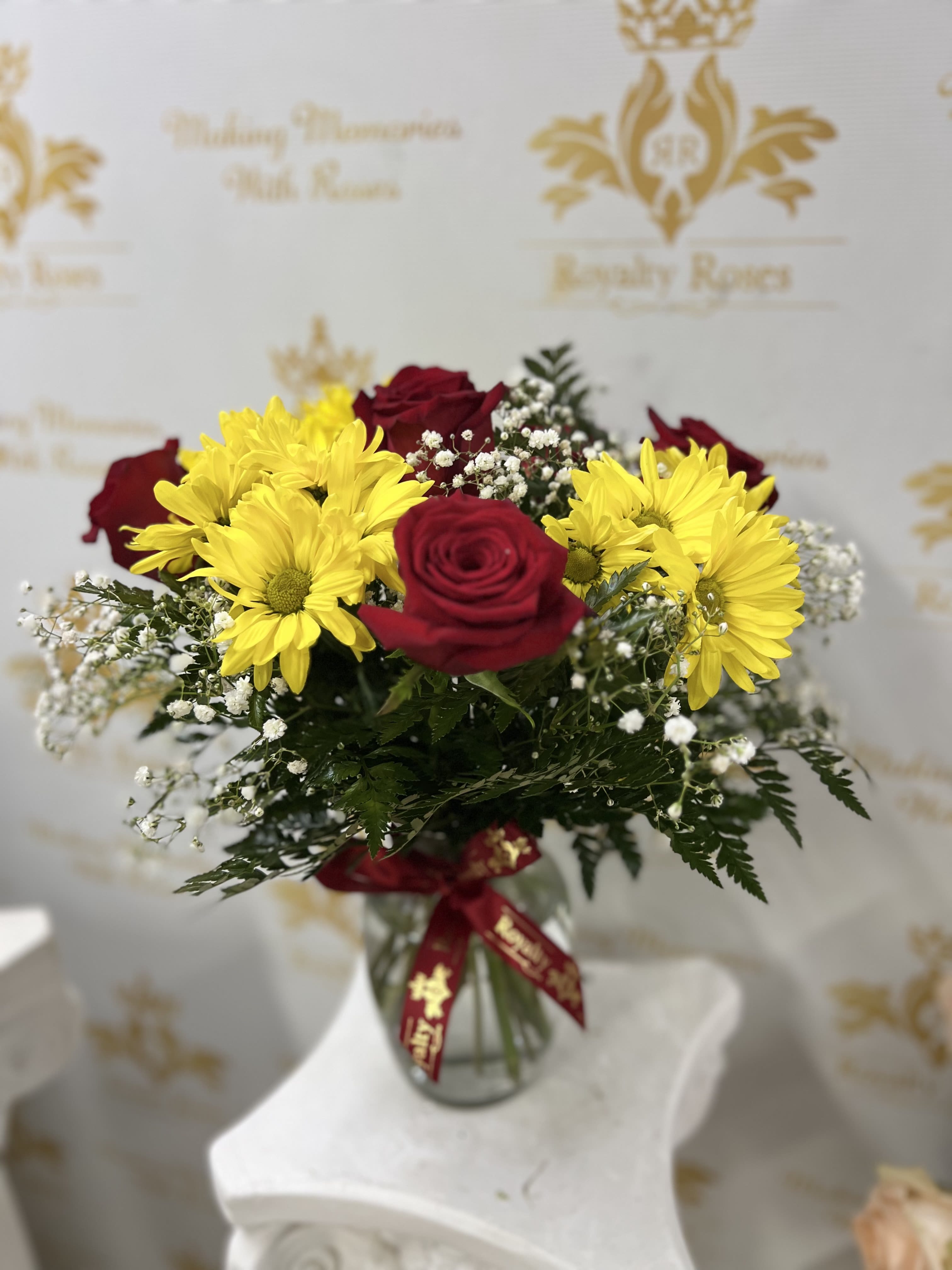 Mothers Days Designers Big Sale - Sunlit yellow daisies paired with rich red roses give this flower arrangement a joyful, romantic energy-perfect as a Mother's Day bouquet, birthday surprise, or anniversary gift. The bright color contrast and airy white filler make the person you're thinking of feel celebrated and cherished the moment they see it.  Two deep red roses sit among cheerful yellow daisy-like blooms and clusters of small white filler flowers, arranged in a rounded, hand-gathered composition. The stems are presented in a clear glass vase and finished with a red satin ribbon tied at the neck for a polished, gift-ready presentation. The mix of bold color, soft texture, and a classic vase makes this arrangement equally suited for everyday gifting, thank-you gestures, or love-themed occasions.  Designed to arrive ready to display, the bouquet measures to fit standard tabletop vases and arrives wrapped with secure padding for safe delivery. To keep blooms fresh longer, change the water every two days and trim stems at an angle before placing in fresh water; gentle daily misting of the filler flowers helps maintain their light, airy look.