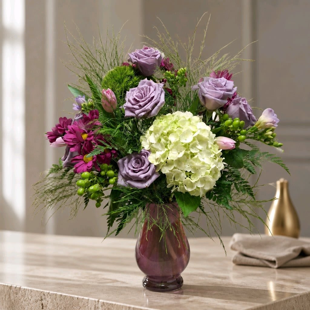 Soft Romance  - Lavender roses and a single voluminous green hydrangea create the soft focal point of this romantic flower arrangement - a perfect birthday bouquet, anniversary roses alternative, or thinking-of-you gift that reads fresh and intimate. Hints of deep magenta daisy-like blooms against airy green foliage give the composition a gentle, garden-style silhouette that will make the person you're sending it to feel truly seen and celebrated.  This rounded arrangement is presented in a pink glass vase that echoes the lavender tones of the roses and adds a polished decorative finish for display. Expect a mix of roses, hydrangea, daisies and fine, airy greenery arranged for a full yet effortless texture in pink, purple, white and green hues. The compact, rounded shape sits beautifully on tabletops and side tables; fill-and-care are the usual steps-fresh water and a clean vase keep blooms looking their best for days-making it an easy, heartfelt gift for everyday celebrations or special moments.