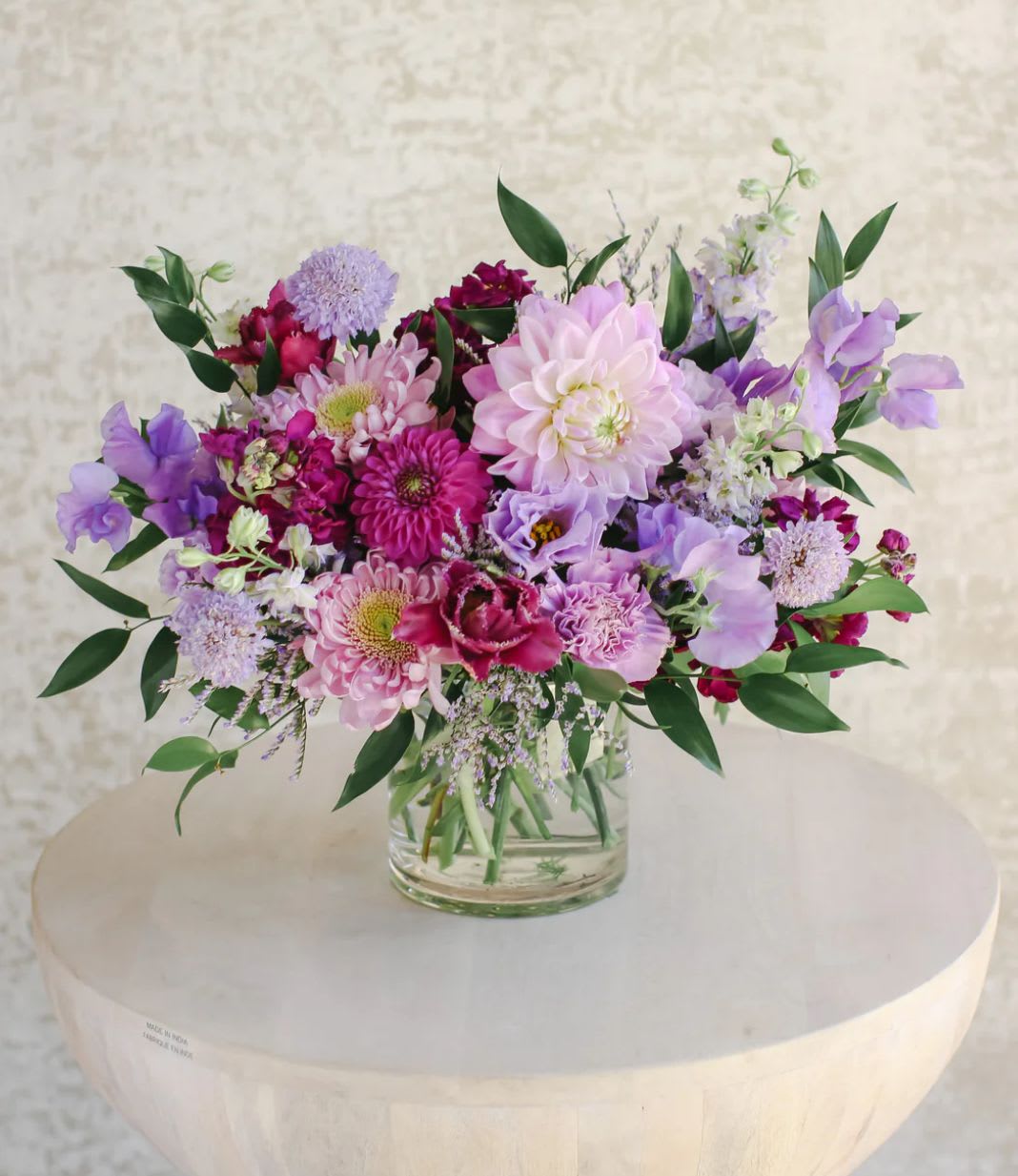 Low glass vase of pink and purple mixed flowers arranged in a lush, rounded centerpiece
