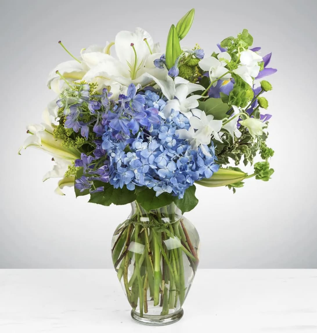 Mixed bouquet of blue hydrangeas, white lilies, and purple blooms in a clear glass vase