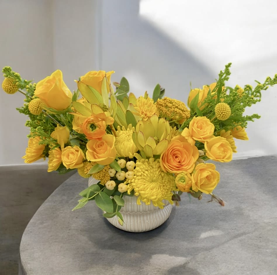 Low yellow flower arrangement with roses and other blooms in a white ceramic pot.