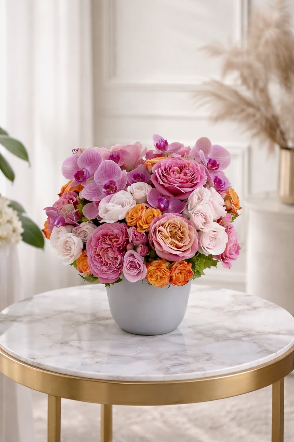 Low round arrangement of pink roses, orchids, and orange spray roses in a white pot on a marble table