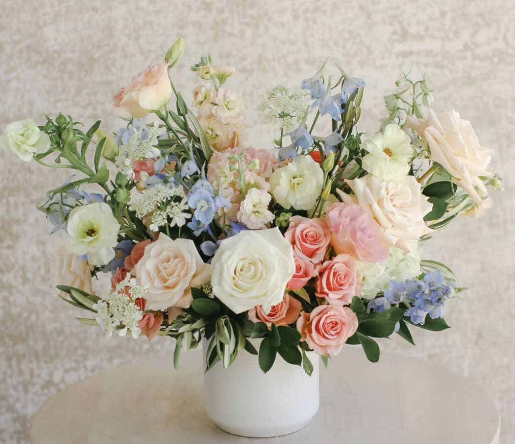 Pastel roses, lisianthus, and blue delphinium in a white ceramic vase on a light table