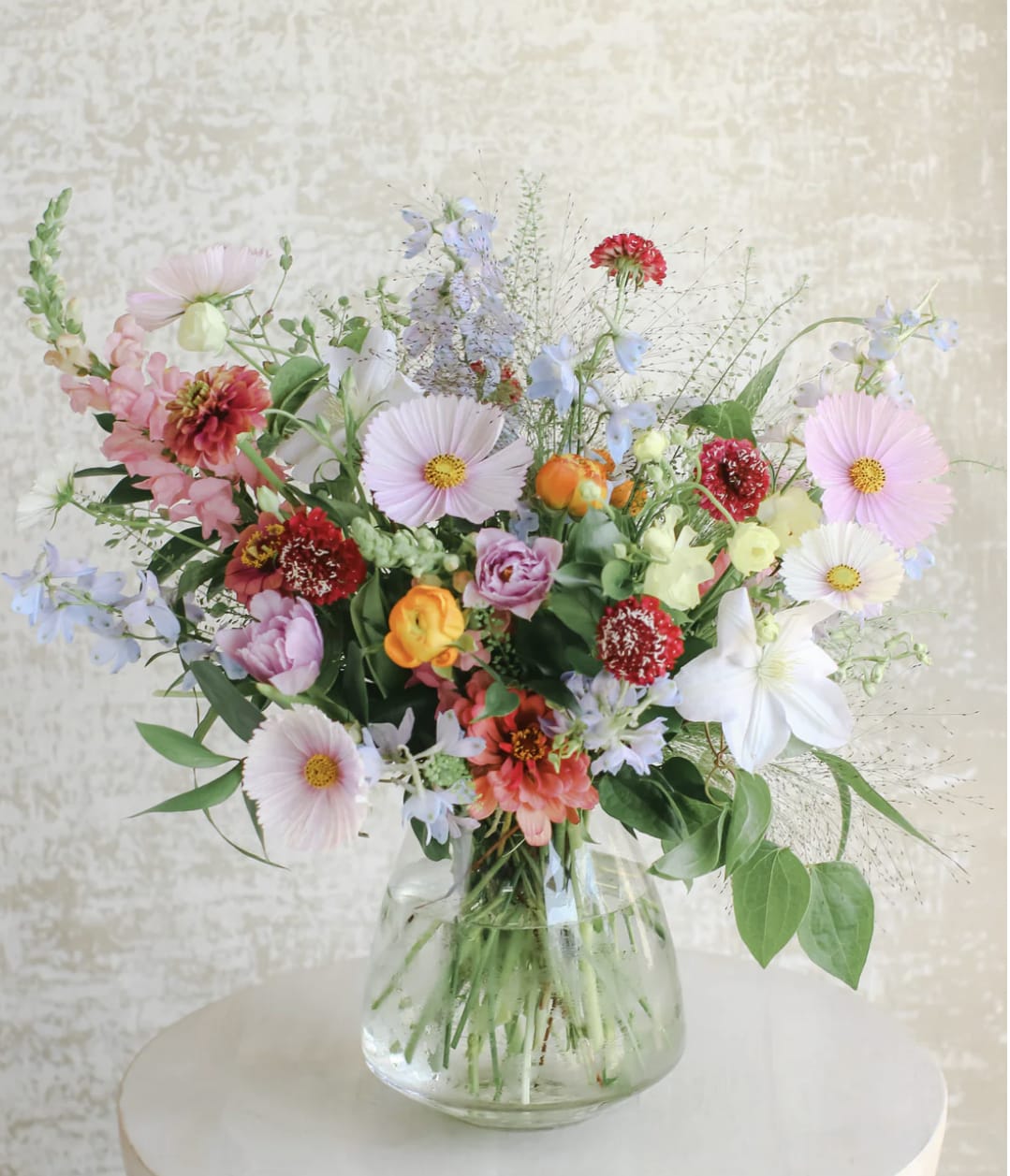 Loose garden-style bouquet of mixed pastel and bright flowers in a clear glass vase.