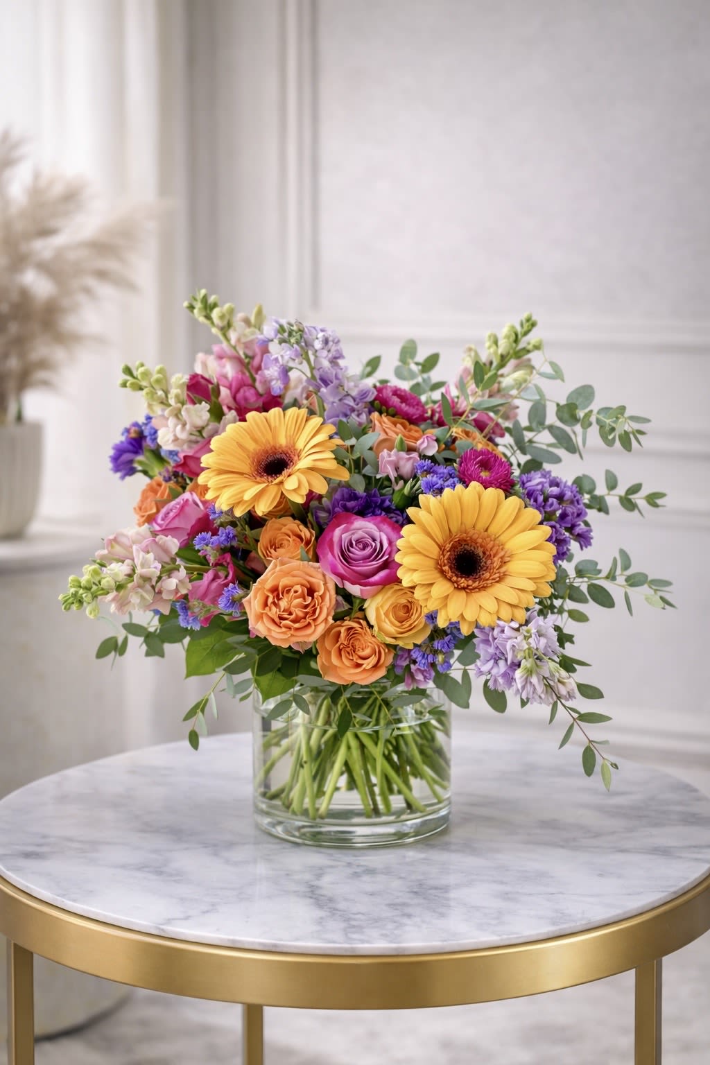 Bright mixed bouquet of yellow gerbera daisies and roses in a clear glass vase on a round table.