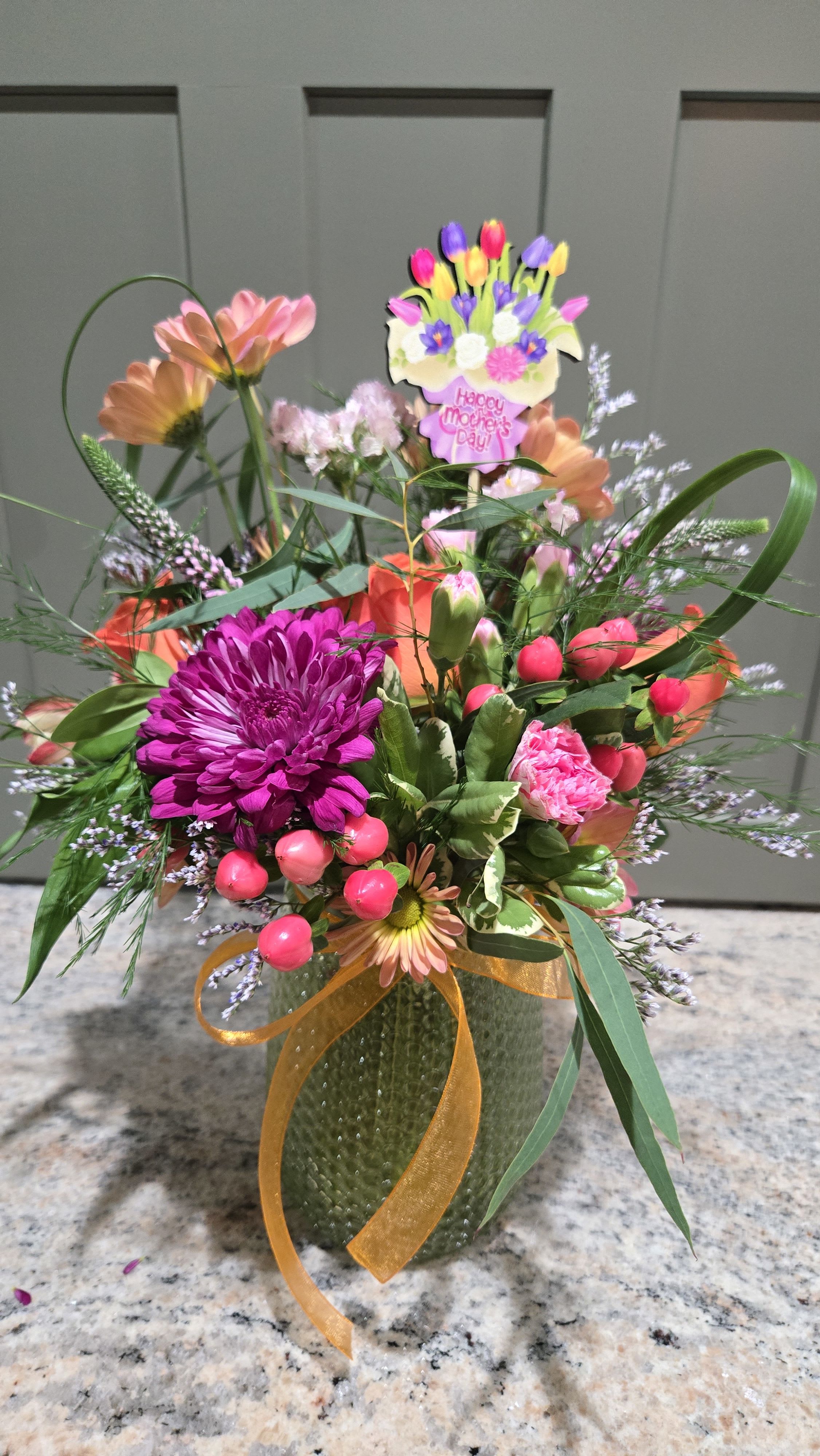 Just For Mum - A vibrant celebration of spring, this flower arrangement is made just for Mom - a cheerful Mother's Day bouquet that brightens her day with pinks, purples, oranges and soft peaches. The colorful mix reads like a sunny garden in bloom, the perfect way to make the person you're thinking of feel seen and celebrated on Mother's Day or any special morning you want to surprise her.  Dense clusters of chrysanthemums, roses, carnations, daisy-style blooms and tulip-shaped flowers mingle with airy filler and greenery for texture and movement. A large magenta chrysanthemum anchors the front while coral and white accents lift the palette; everything is arranged in a textured green glass vase and tied with a sheer orange ribbon. A decorative "Happy Mother's Day" pick tops the design, giving it a festive, gift-ready finish that arrives ready to display.