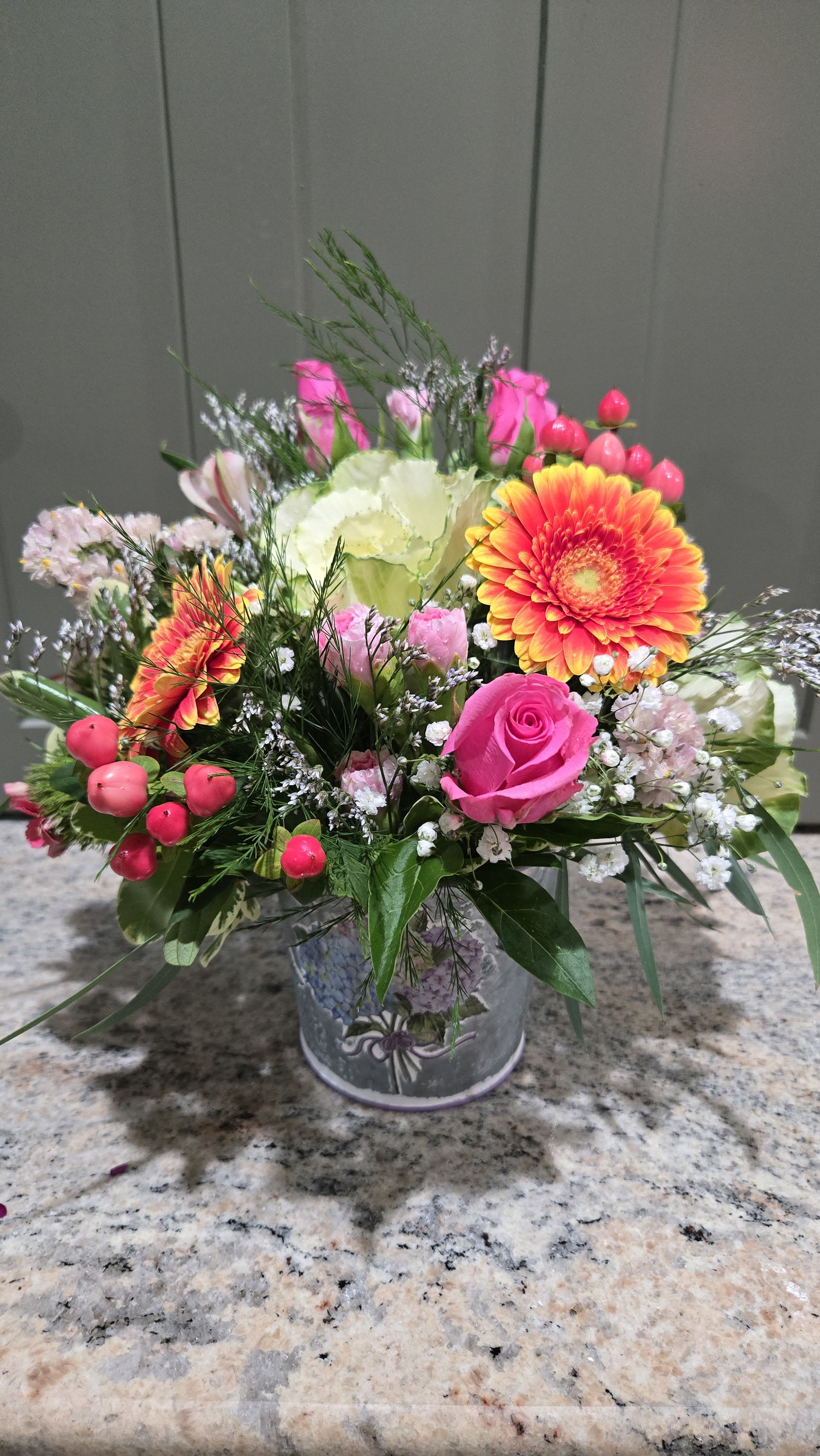 Petite, But Bold - Vivid pink roses and sunny orange-and-yellow gerbera daisies make a joyful statement in this petite but bold flower arrangement-perfect as a birthday bouquet, a cheerful thank-you gift, or a bright spring centerpiece. The color story of hot pink, warm orange, soft white, and fresh green announces celebration at first glance, so the person you're thinking of will feel noticed and uplifted the moment it arrives.  The low metal container holds a rounded, full design of roses, gerbera daisies, pale carnations, and small white filler blooms, accented with airy greenery and berry-like stems for texture and movement. Compact yet abundant, the arrangement sits low and wide for easy placement on a table, desk, or countertop and brings lively color without overwhelming a space.  Ideal for friends, coworkers, or anyone who appreciates bold color in a modest package, this arrangement arrives arranged and ready to display. For best enjoyment, keep the flowers in a cool spot away from direct heat and refresh the water daily to extend their vase life.
