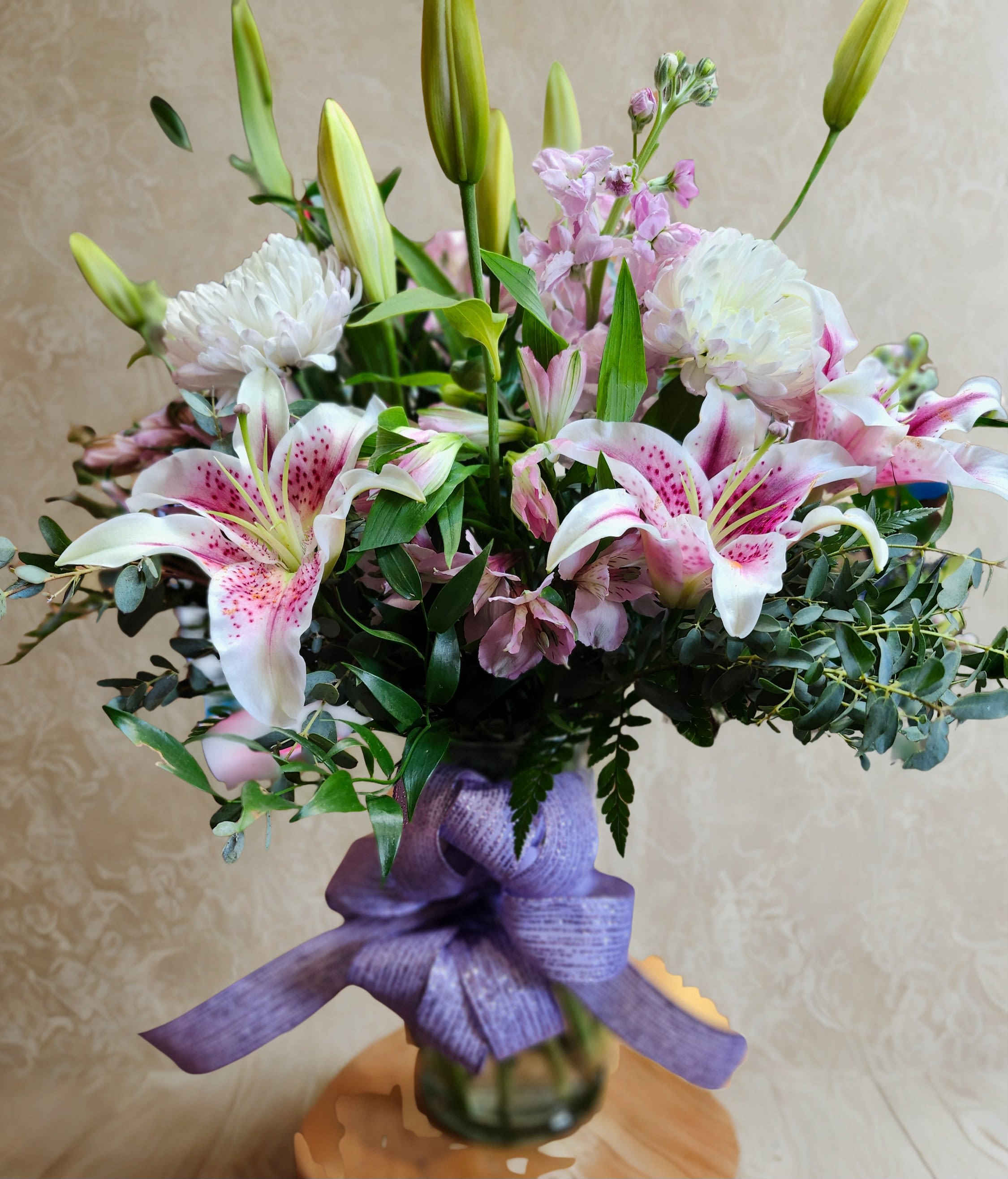 Lily Elegance - Soft, graceful, and effortlessly sophisticated, Lily Elegance is a stunning arrangement designed to make a lasting impression. Featuring fragrant stargazer lilies surrounded by delicate pink blooms, crisp white mums, and fresh eucalyptus, this bouquet blends romantic colors with lush textures for a look that feels both timeless and modern. Arranged in a clear glass vase and finished with a flowing lavender ribbon, it’s the perfect choice for birthdays, anniversaries, celebrations, or simply brightening someone’s day with beauty and love.