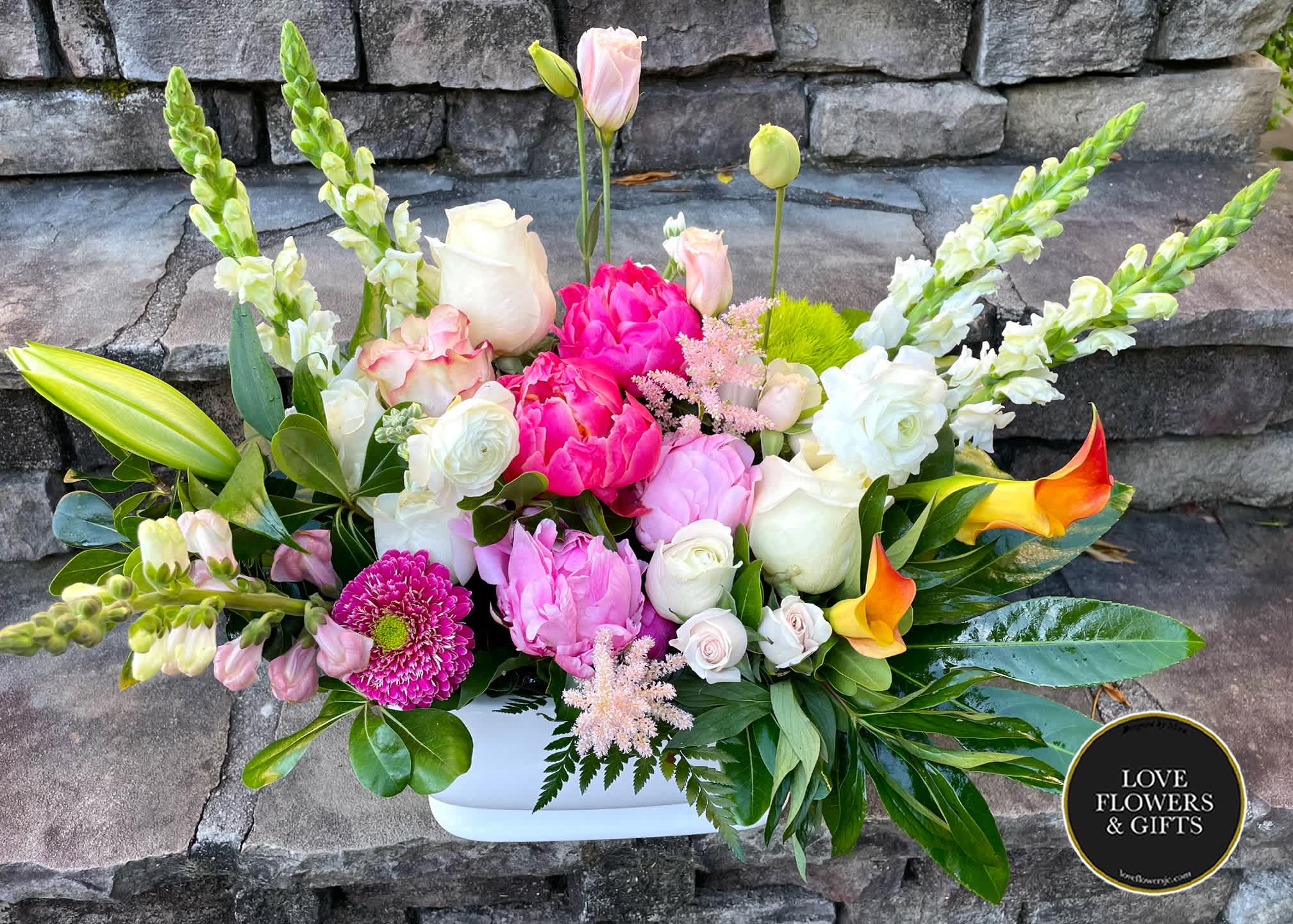 Rectangular arrangement of pink peonies, white roses, snapdragons and orange calla lilies in a white container.