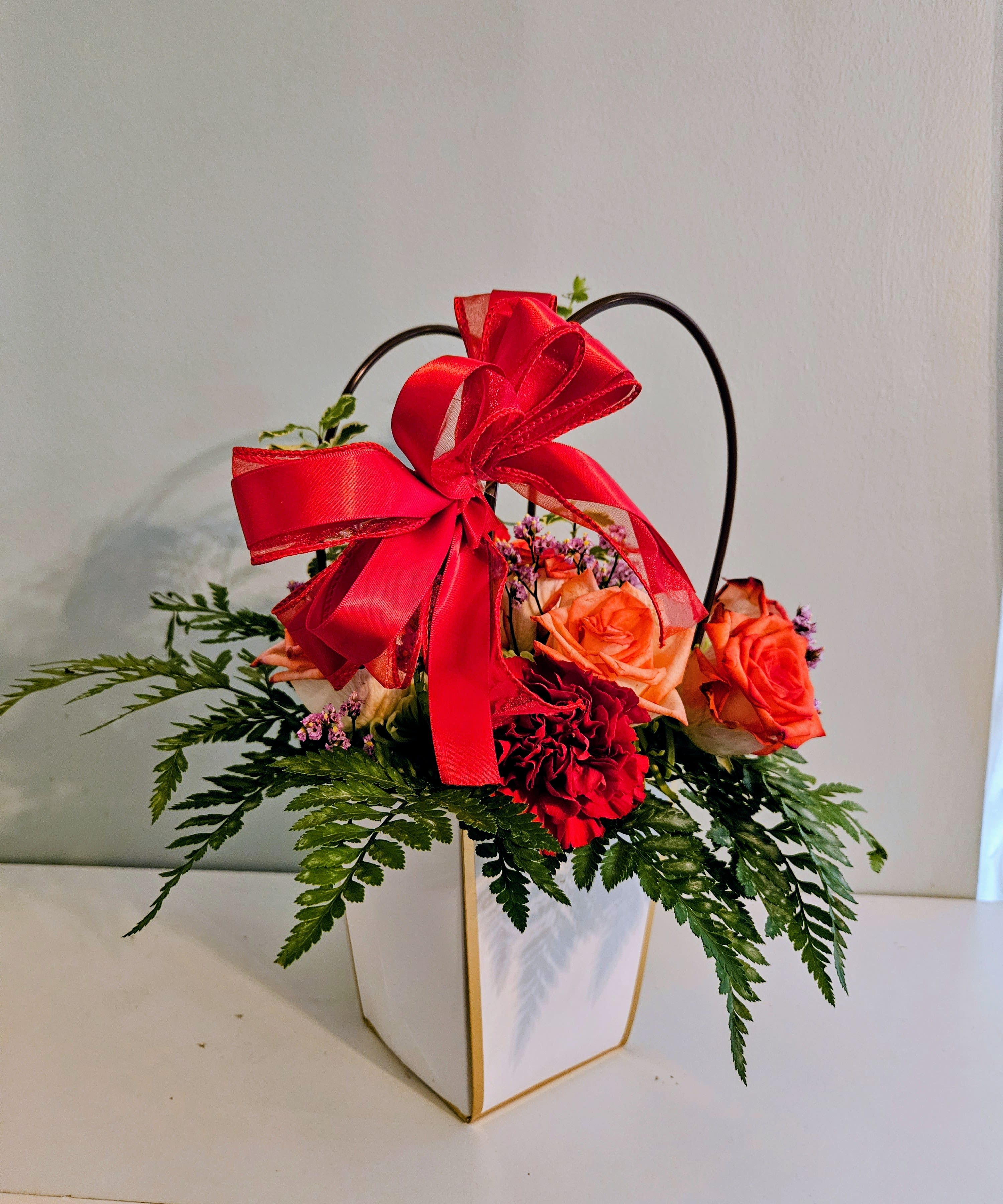 "Mother`s Day" Beauty Flower Mix Bag - Bright, warm and gift-ready, this Mother's Day flower arrangement mixes roses, carnations, lilies and cheerful daisy-like blooms in a compact box perfect for a birthday, anniversary, or Mother's Day surprise. Presented in a white paper gift box with a black arched handle and a bold red satin bow (other options show pink or gold-trimmed boxes with matching ribbon), the design reads as celebratory and polished-she'll feel truly seen when the ribbon is untied and the layered bouquet is revealed.  The mix features orange and red roses, deep red carnations, pink filler flowers, white lilies, small purple and white daisy-style accents, and fern-like greenery that spills over the edges for a full, textured look. Sizes are compact and basket-style, arranged in rectangular gift boxes or geometric baskets with woven or black handles; each comes finished with a large decorative ribbon bow for an immediate "gift" presentation. The color story-reds, oranges, pinks, greens, whites and purples-creates a warm, joyful impression that suits romantic moments, thank-you gestures, and celebratory occasions alike.