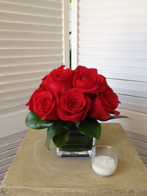 Red roses arranged in a square glass vase with a small candle nearby
