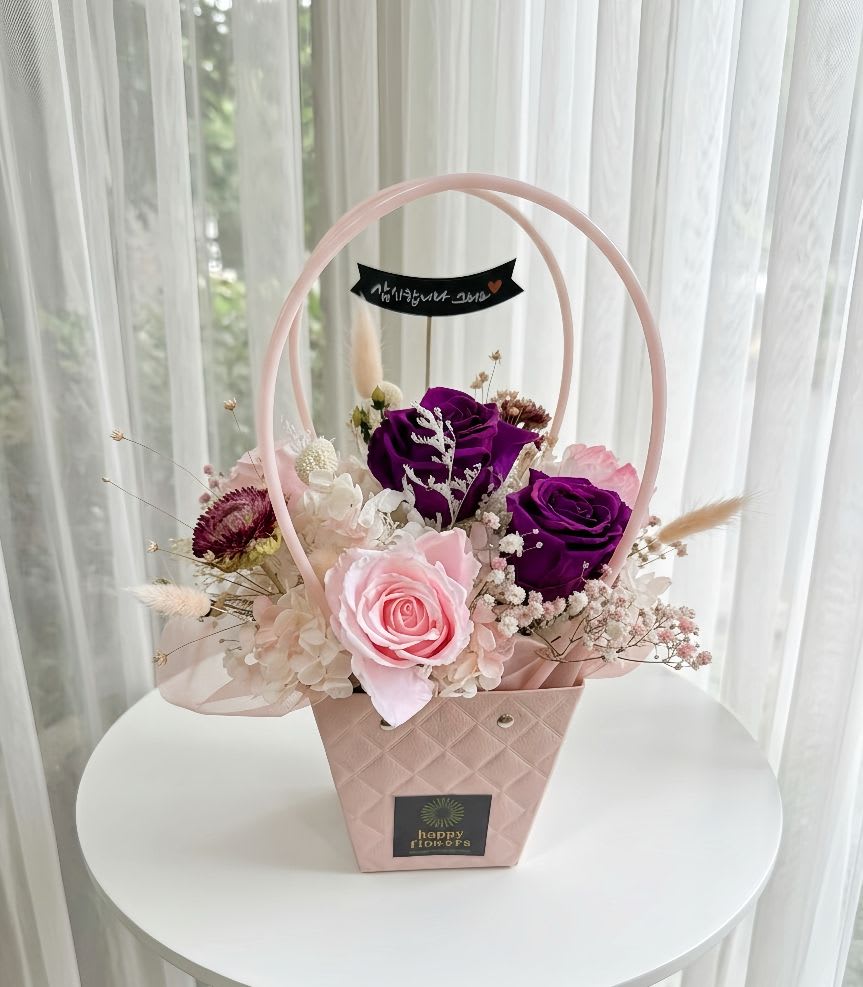 Preserved Flower Gift Baskat #1 - A charming preserved flower arrangement featuring 2 premium deep purple roses and 1 soft pink rose. This elegant bouquet is accented with white hydrangeas, pink baby's breath, dried strawflowers, and fluffy bunny tails, all beautifully displayed in a stylish pink quilted-texture gift basket with circular handles. 