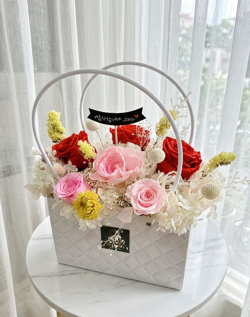 Preserved Flower Gift Baskat #2 - A vibrant preserved flower arrangement featuring 3 bold red roses and 3 soft pink roses. Nestled in a bed of white hydrangeas, this elegant piece is accented with yellow Billy balls, white baby's breath, and dried floral textures, all beautifully presented in a white quilted-texture gift bag with circular handles.