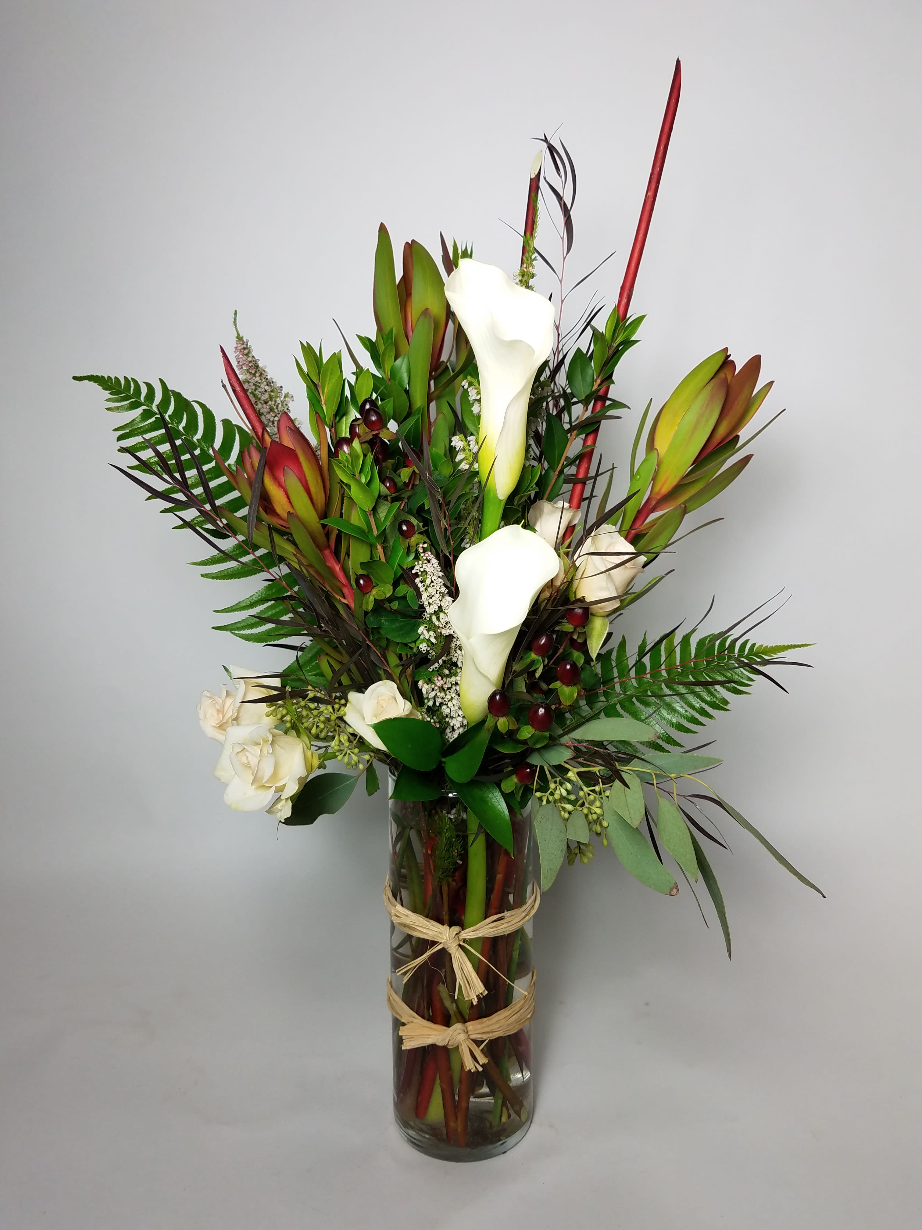 Tall arrangement of white calla lilies and red tropical blooms in a glass vase