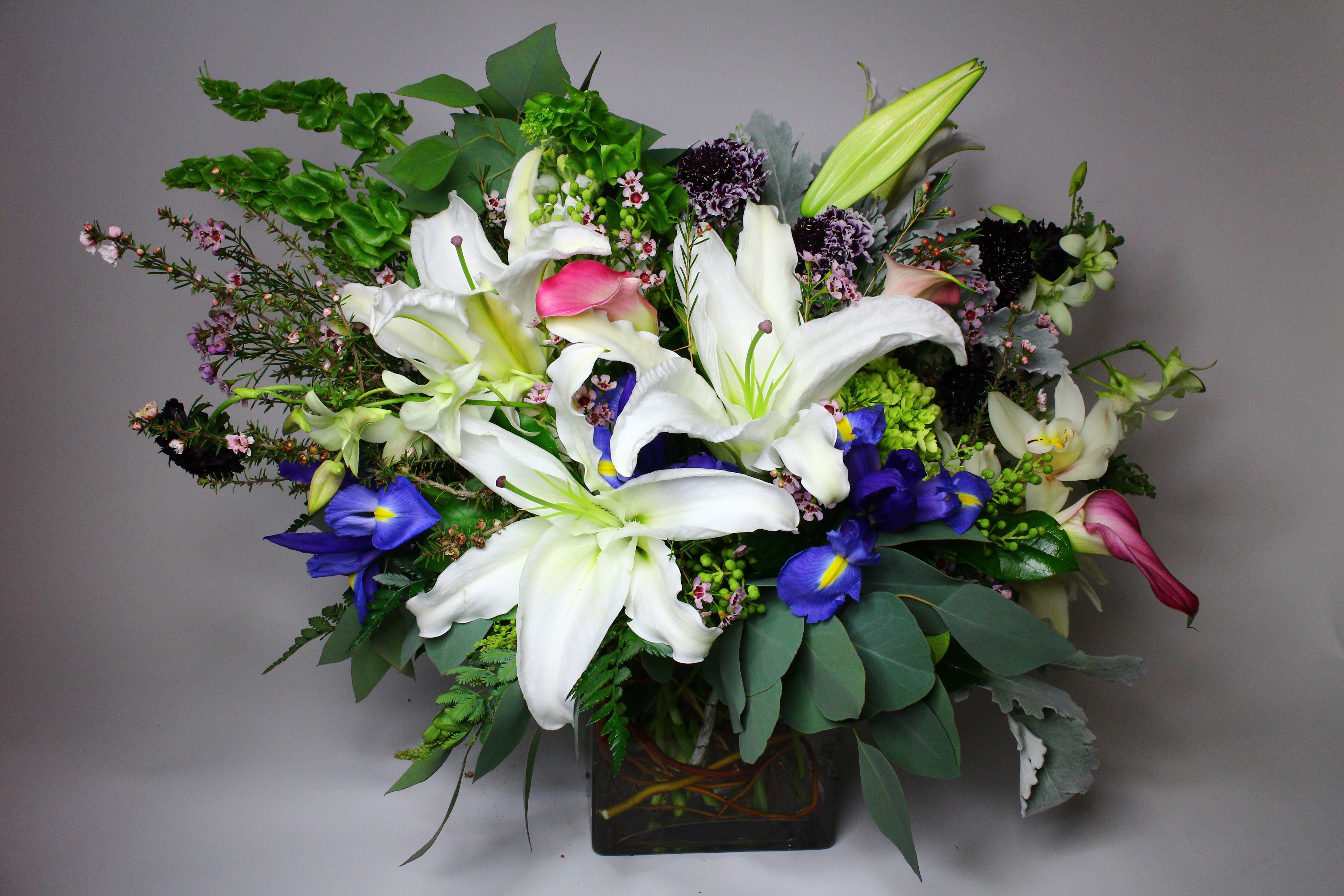 Mixed bouquet with white lilies, blue irises, and pink accents in a square vase