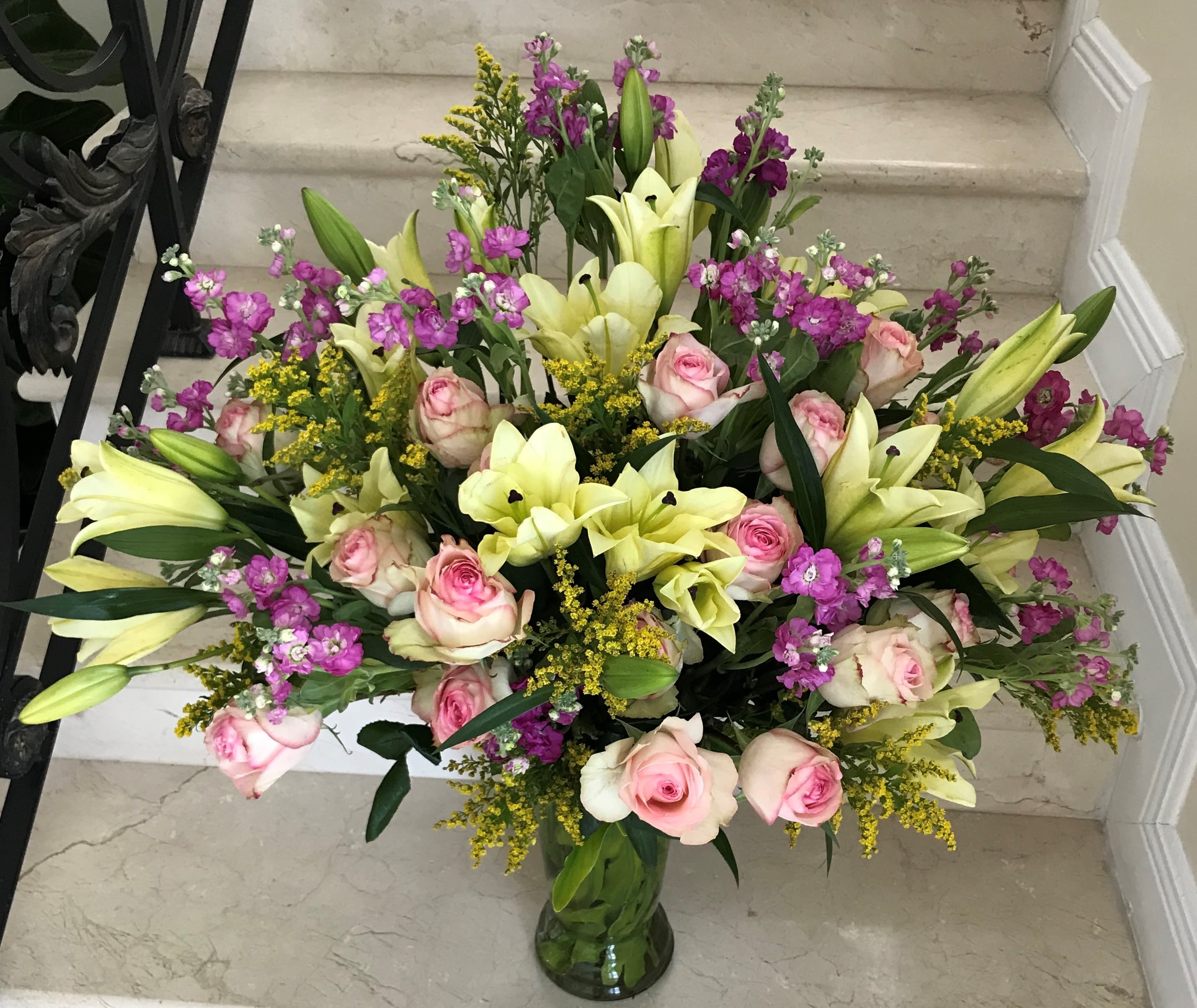 Large mixed bouquet of pale yellow lilies, pink roses, and purple flowers in a clear glass vase