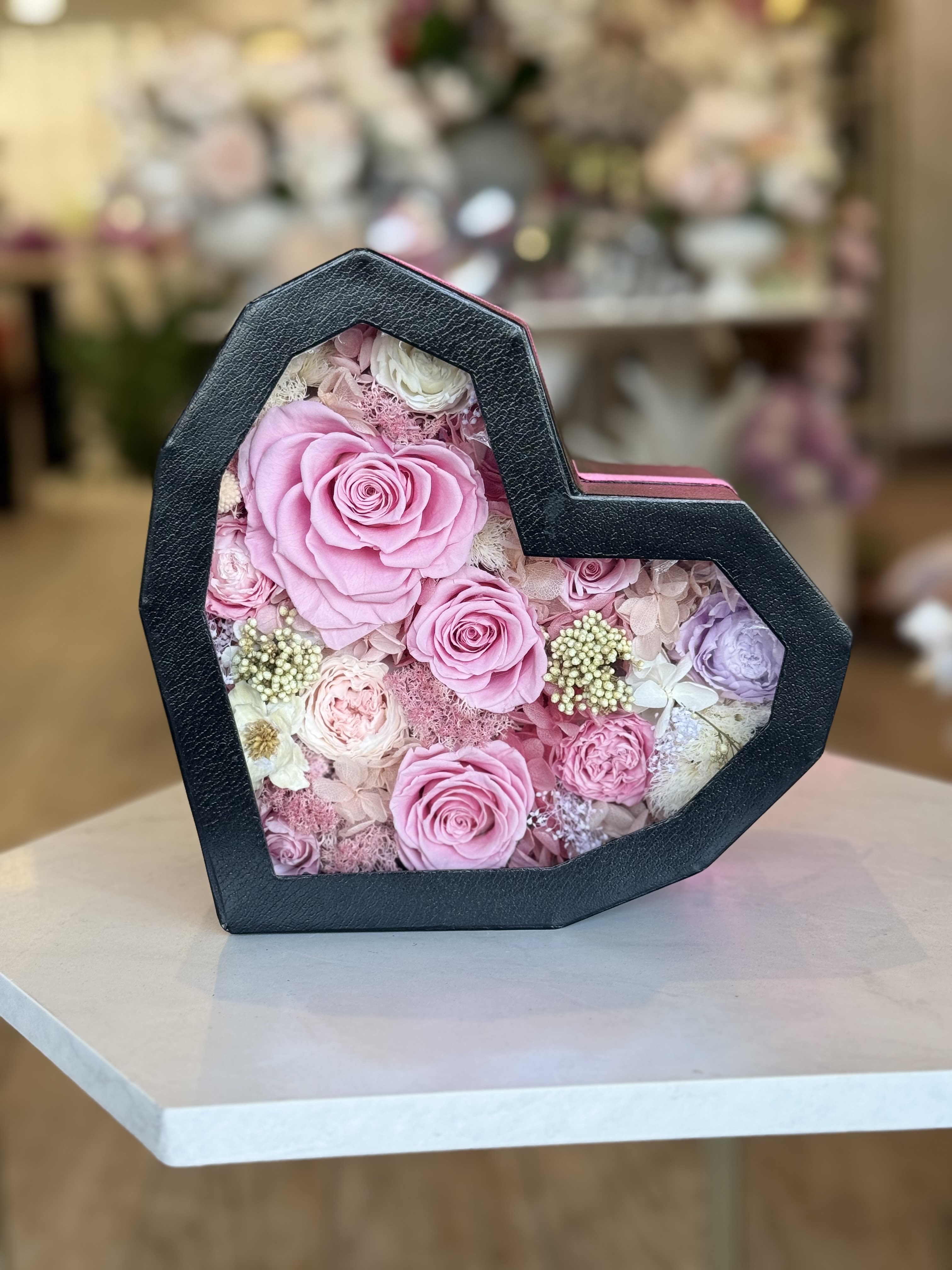 Beautiful preserved flowers and necklace in heart box  - A heart-shaped black gift box reveals a keepsake of preserved roses and hydrangea accents under a clear lid-an intimate flower arrangement perfect for an anniversary, birthday, Mother's Day, or a romantic "just because" surprise. The pink ribbon and included heart pendant necklace tuck into a lower compartment so the recipient opens both flowers and a small treasure; the presentation reads like a moment made to be unwrapped and remembered.  Tightly packed blush, pale pink, lavender and ivory roses mingle with soft dried hydrangea and delicate filler blooms for a layered, textured look that feels both fresh and everlasting. The compact, layered surface measures as a gift-box style arrangement (heart-shaped black box with window front) that travels well and doubles as a decorative keepsake long after the petals are admired.  Send this when you want her to feel seen and loved-whether it's Valentine's Day, an anniversary, a birthday, or simply to say "I'm thinking of you." No water is needed for the preserved flowers; order options typically include a ribbon and pendant as shown, making it ready to present straight from the box.