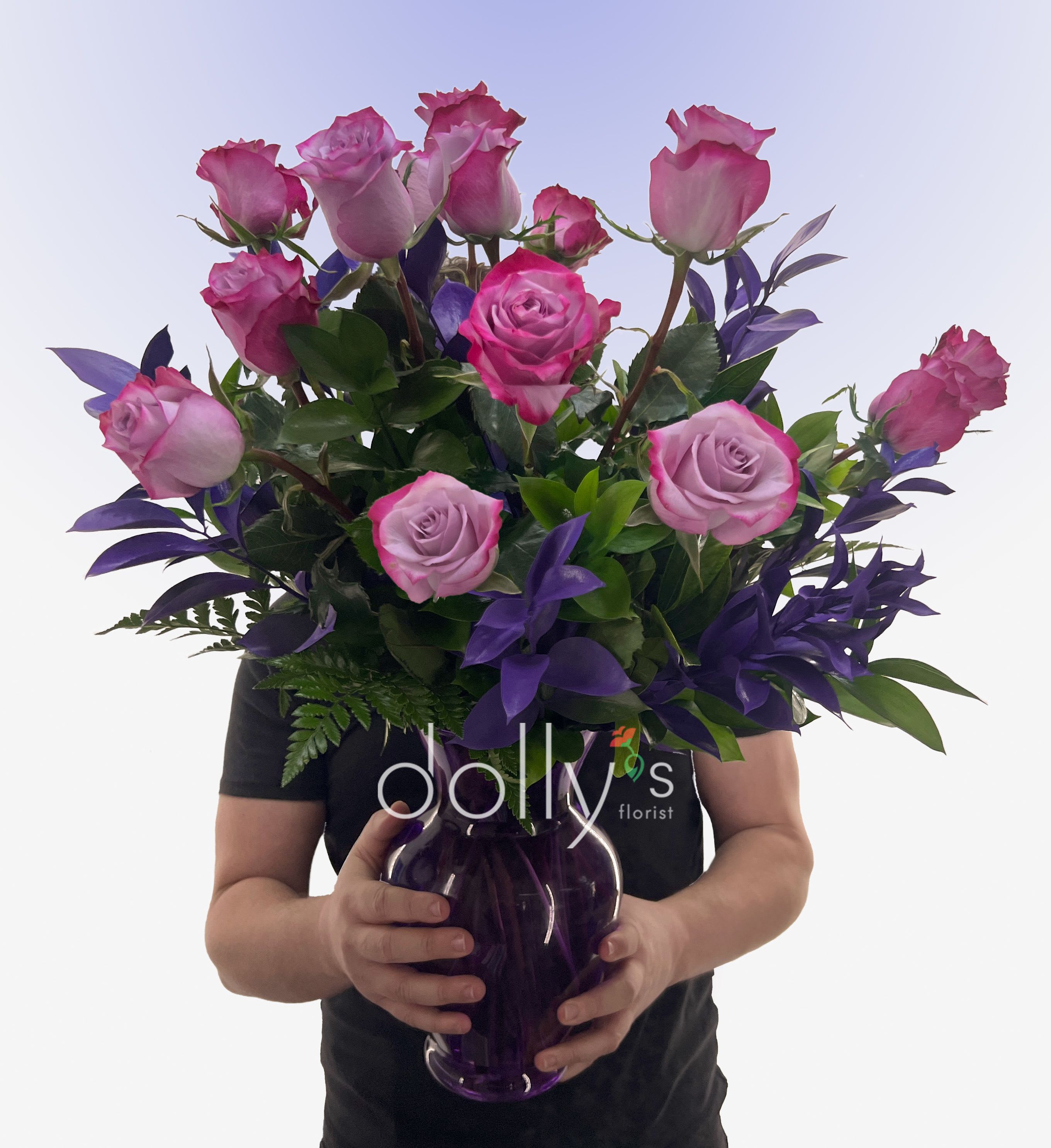 Classic Magenta Long-Stem Rose Bouquet - A dozen magenta long-stem roses arranged in a deep purple glass vase creates an immediate, joyful impression the person you're thinking of will notice - perfect as an anniversary bouquet or a bold "I love you" gesture. The mix of hot pink and softer lavender-pink roses reads both romantic and modern, making this arrangement equally right for Valentine's Day, a congratulations gift, or a thoughtful "get well" or "thinking of you" surprise.  The roses are clustered in a rounded, abundant shape with rich green leaves and dark purple accent foliage woven between stems to add contrast and depth. The glossy purple vase echoes the arrangement's cooler tones and gives the whole presentation a polished, keepsake quality; dimensions and vase size are chosen to support the long stems and keep blooms upright for a refined silhouette.   Send this to celebrate new baby news, say "I'm sorry" with sincerity, or simply to thank someone in a way that feels seen - she'll feel genuinely celebrated when these vibrant roses arrive. For best results, keep the arrangement in fresh water, away from direct heat, and refresh the water every couple of days to extend bloom life.