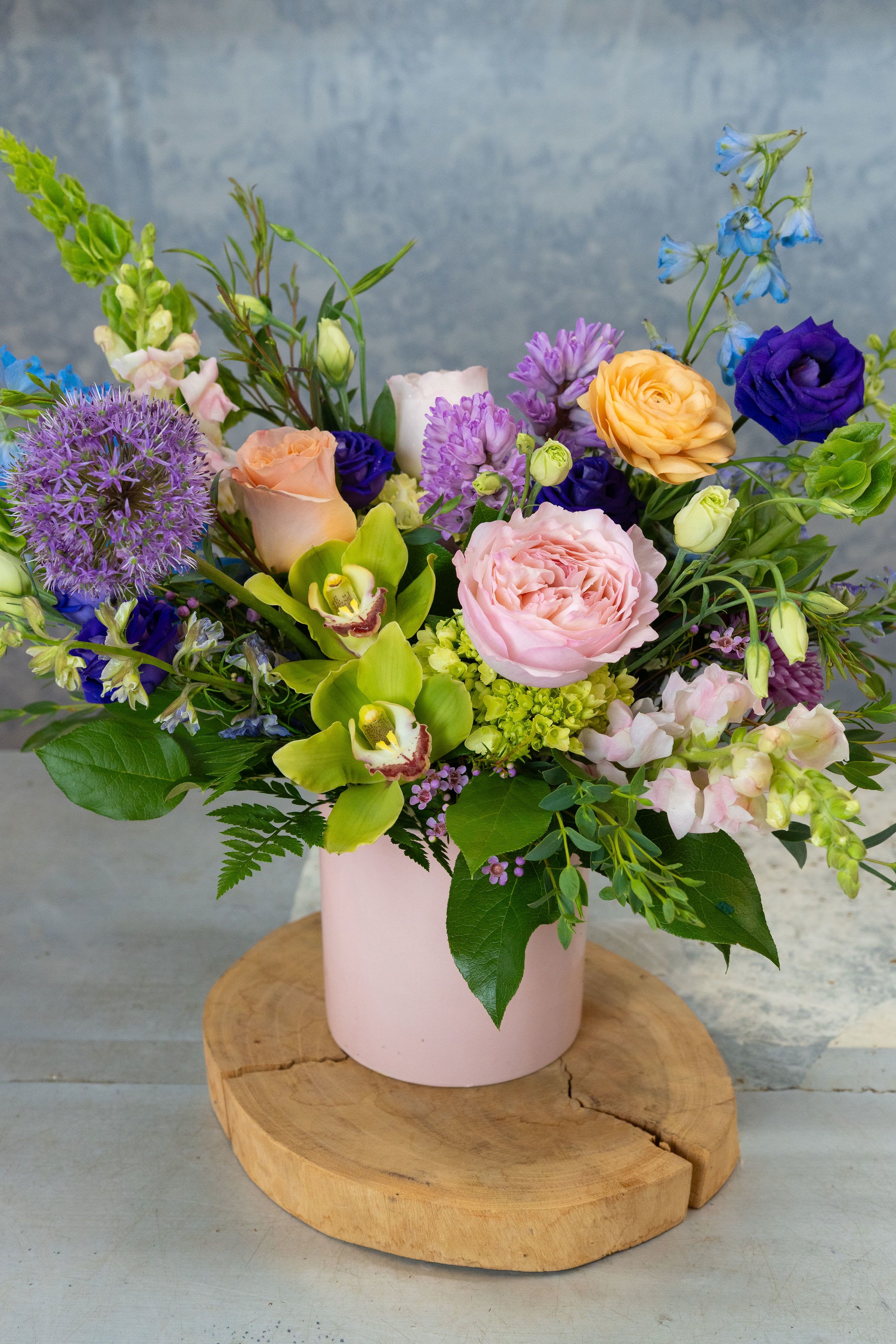 Sunday Brunch - A lush, garden-style flower arrangement that feels like a bright spring morning-perfect as a Mother's Day gift, birthday bouquet, or anniversary surprise. This low, wide mixed arrangement arrives in a soft pink cylindrical vase set on a wooden slab, combining romantic blush and peach roses with vivid purple accents and pops of yellow-green orchids so the person you're thinking of will feel seen and celebrated. The overall effect is lively yet refined, a cheerful centerpiece that reads as both modern and timeless.  Bloom details include hydrangeas, roses, orchids, snapdragons, lisianthus, chrysanthemums, delphinium, allium and other seasonal filler for layered texture: rounded rose heads, airy snapdragon spikes, blue delphinium accents, and a striking purple allium for contrast. Colors range from pastel pinks and peaches to saturated purples, bright yellow-greens and soft blues, arranged compactly in a matte pink vase for easy display on a dining table or desk.  Ideal for spring occasions, birthdays, anniversaries, or a congratulatory gesture, this arrangement is made to brighten a room and lift someone's spirits. It arrives ready-to-display in its pink vase; to keep blooms fresh, place in a cool spot away from direct sunlight and refresh the water every 2-3 days.