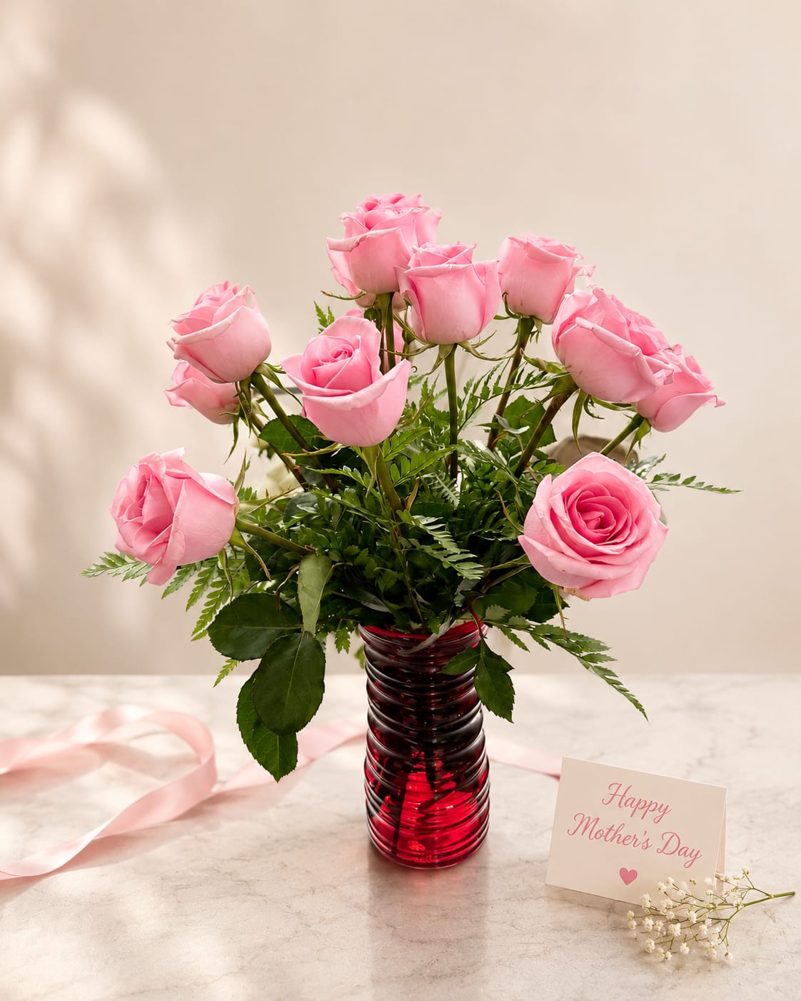 Moms Blush Rose  - Soft pink roses gathered in a ribbed red glass vase create a tender, celebratory bloom perfect for a Mother's Day bouquet or a special birthday surprise. This flower arrangement combines pink, red, white, and green tones to read as both romantic and warmly familial-the kind of gift that makes the person you're thinking of feel seen and cherished.  The arrangement features several open and semi-open pink roses arranged with fern-like filler and glossy rose foliage for an airy, full silhouette. Accents of white and deeper red tones appear in the mix, and a pink ribbon ties the vase for an extra touch of sweetness. It arrives in a striking ribbed red glass vase that adds height and contrast while keeping stems neatly gathered.  Choose this as a springtime gift for mom, grandma, or any mother-figure to mark Mother's Day, a birthday, or "just because" moments. A small greeting card reading "Happy Mother's Day" is included with the presentation; standard care is to refresh vase water every two days and trim stems slightly to extend bloom life.