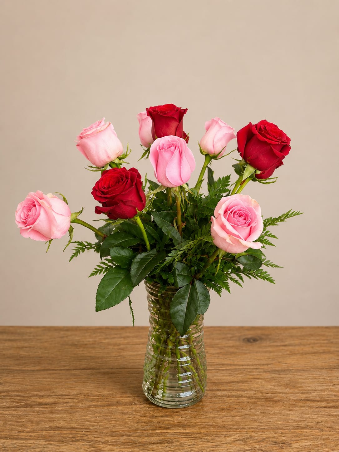 Classic Rose Bloomnation  - A loose cluster of pink and red roses arranged at varying heights creates an airy, romantic centerpiece that reads classic and effortless-the perfect rose arrangement for an anniversary, Valentine's Day, or a thoughtful everyday gift. The mix of soft pink and saturated red roses sends a clear message of affection and warmth so the person you're thinking of will feel cherished the moment these blooms arrive.  The bouquet sits in a clear ribbed glass vase that showcases dark green foliage filling the lower half for clean contrast against the petals. Blooms are arranged upright with natural spacing for a relaxed, garden-picked look; the combination of color, texture, and height gives the display elegant movement without feeling fussy.  Ideal for dining tables, bedside tables, or an office desk, this arrangement makes someone feel genuinely celebrated whether it's a milestone or just because. Vase-ready and low-maintenance-replace water every few days and trim stems if needed-to keep the roses looking fresh throughout their vase life.