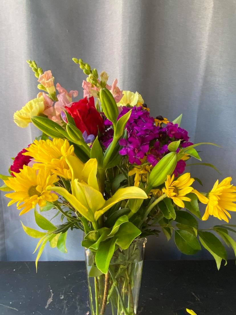 Mixed bouquet of yellow daisies, lilies, and purple flowers in a clear vase