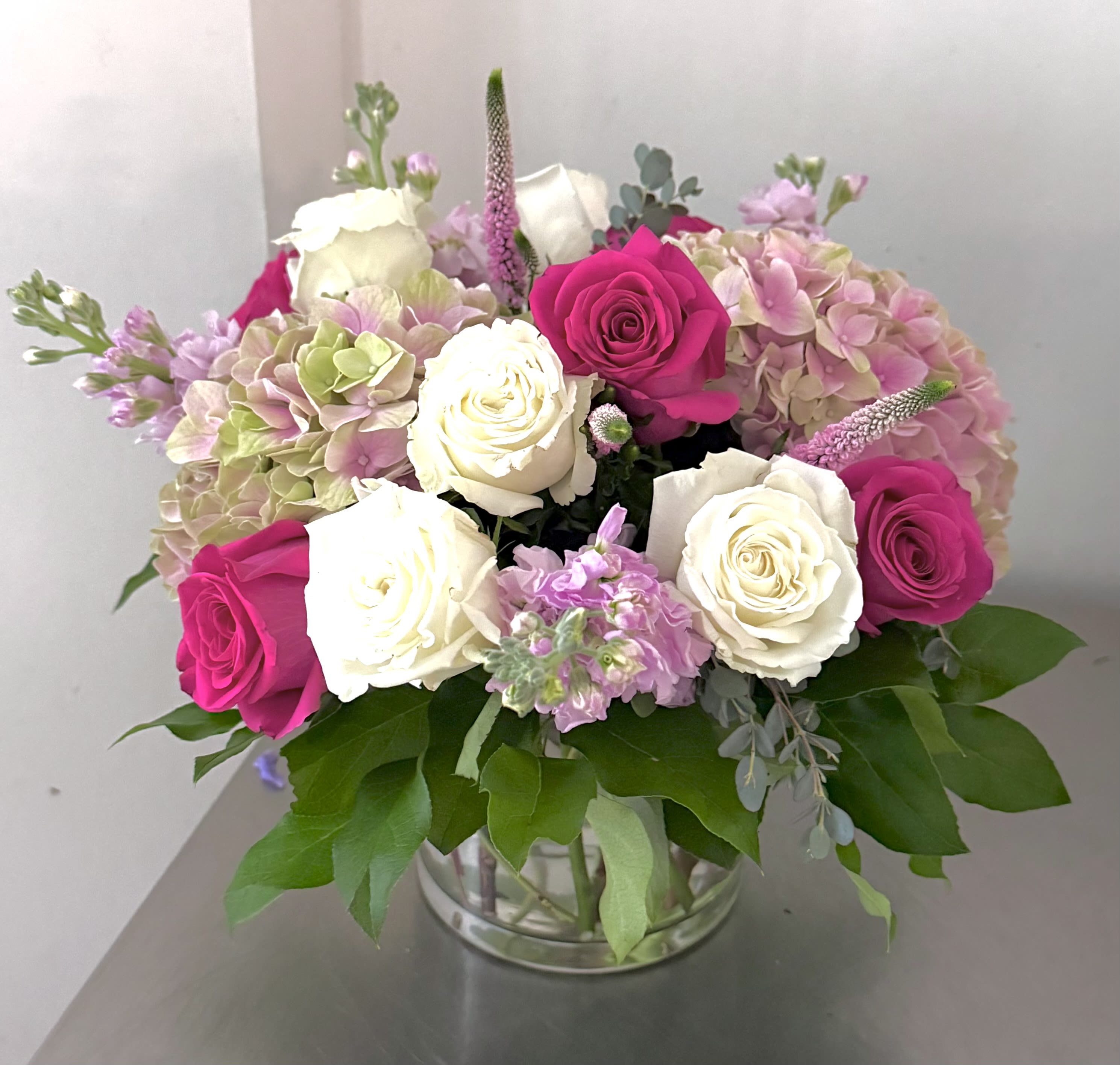 Blush &amp; Berry Garden Rose Arrangement - A romantic, garden-inspired arrangement designed in a low clear glass vase, featuring lush pink hydrangea, elegant white and fuchsia roses, delicate stock, and soft textural accents. Layers of fresh greenery frame the blooms, creating a full, airy composition that feels both refined and naturally abundant.  This design blends classic roses with seasonal garden florals in shades of blush, berry, and ivory—perfect for birthdays, anniversaries, thank-you gifts, or simply brightening someone’s day with timeless beauty.  Handcrafted with premium fresh flowers and arranged to showcase dimension from every angle, this piece brings a soft, graceful presence to any table or home setting.