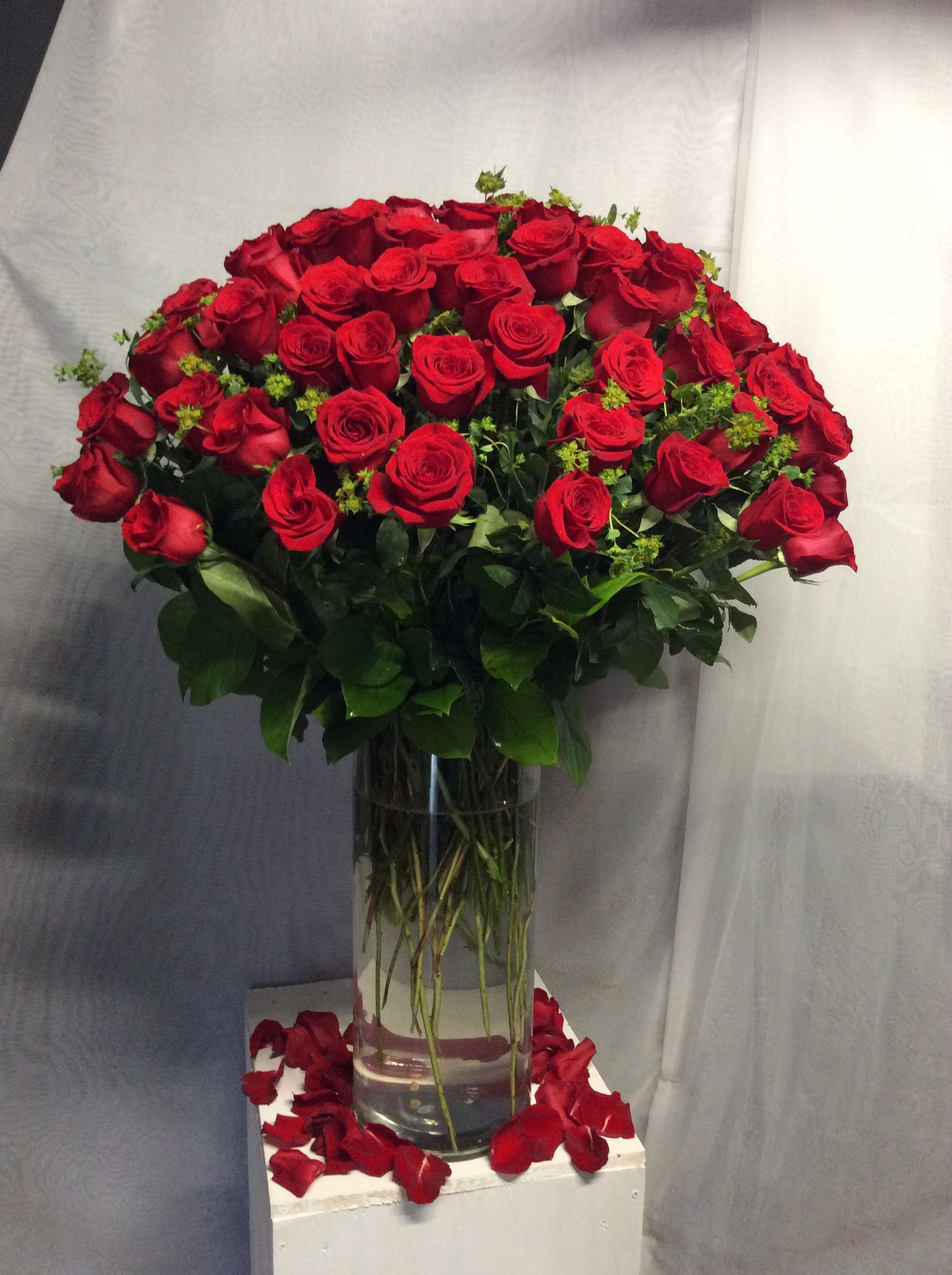 Large arrangement of long-stemmed red roses in a tall clear glass vase with petals scattered below.