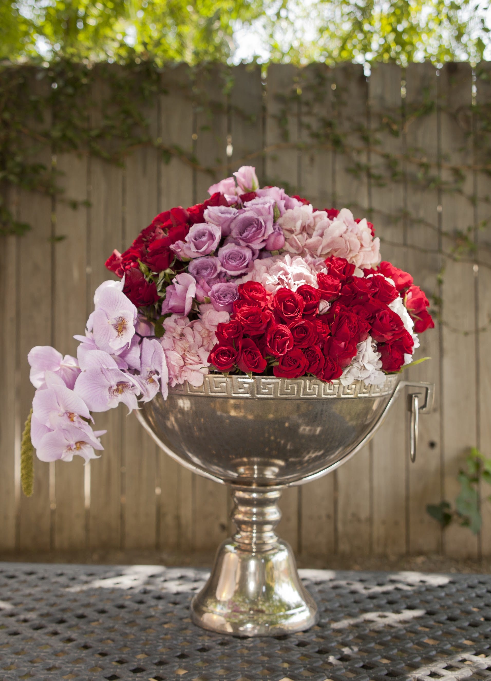 Pink, red, and lavender roses arranged in a silver urn with orchids draping over the side