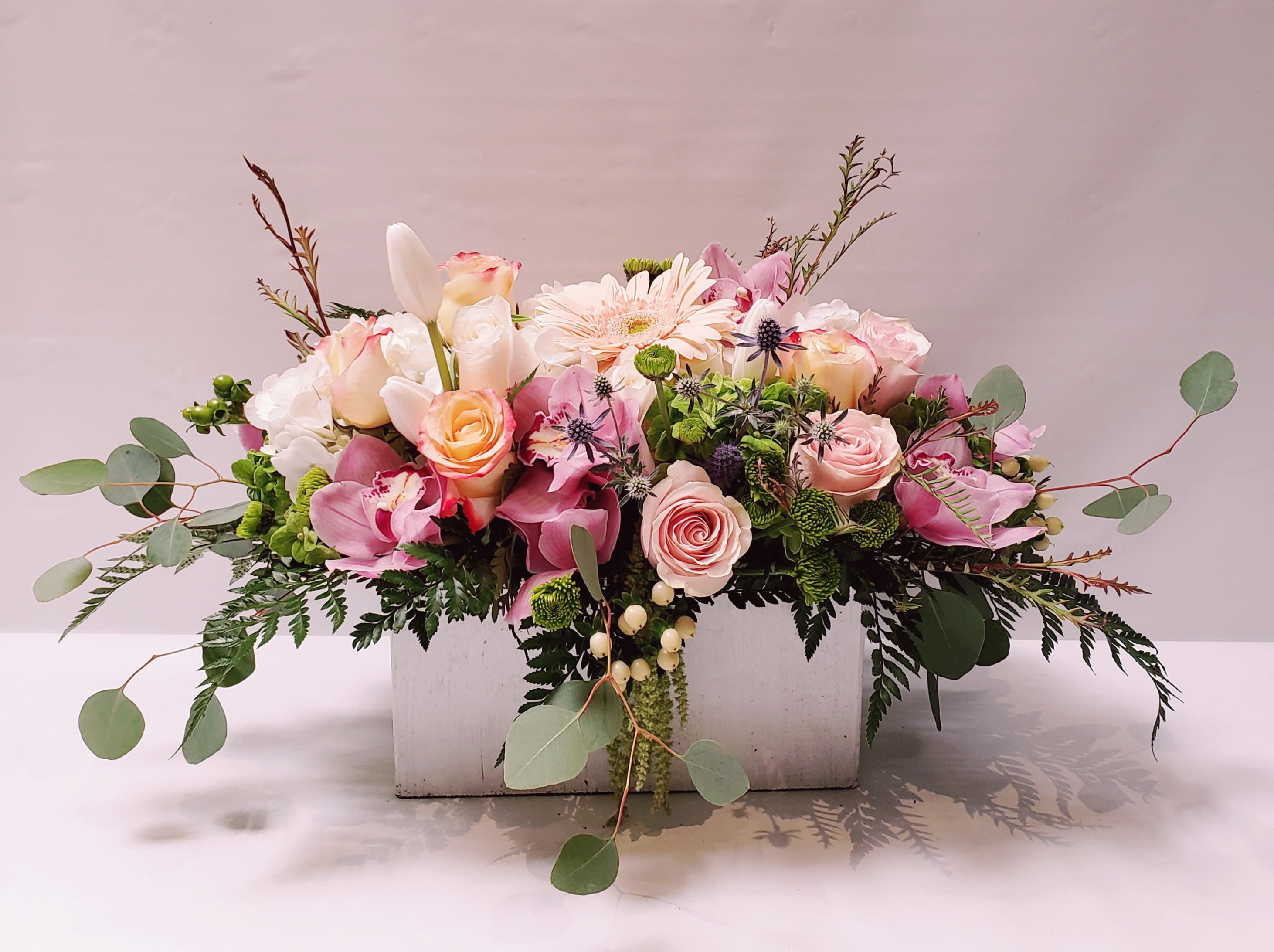 Low pastel arrangement of roses, orchids, and mixed blooms in a white wooden box.