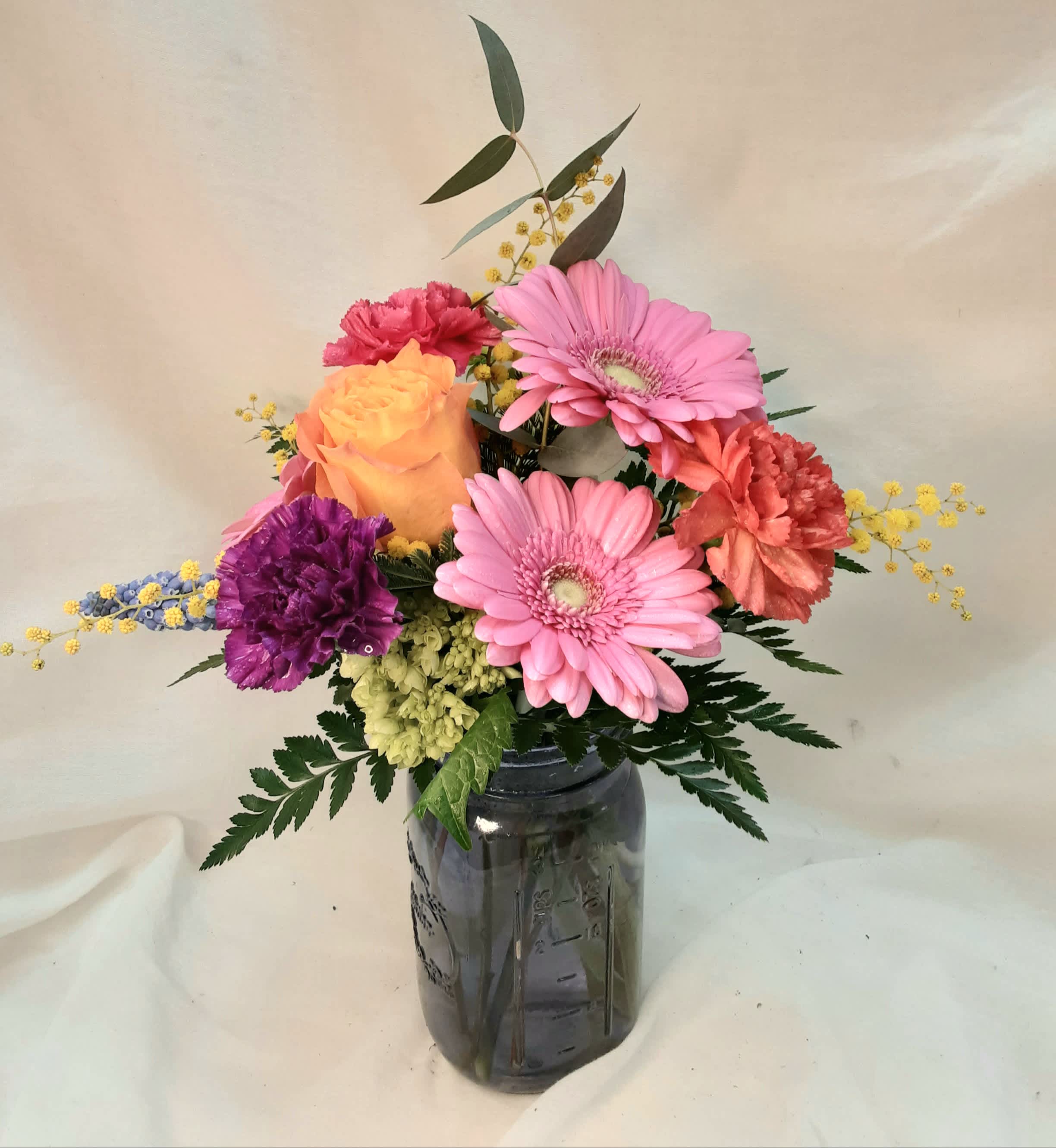 Colorful mixed arrangement of pink gerbera daisies, orange carnations, and yellow rose in a tinted glass jar